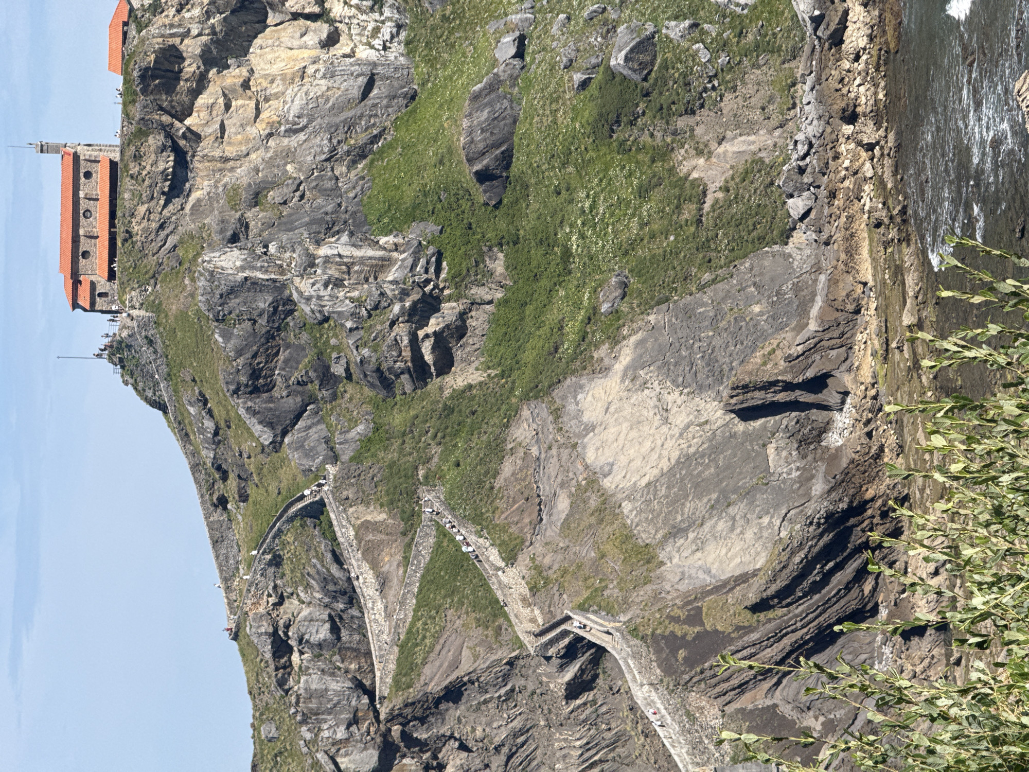 The image depicts the San Juan de Gaztelugatxe hermitage, a historic religious site located off the coast of Bermeo, Spain. A winding stone staircase, consisting of approximately 241 steps, ascends a rocky islet, connecting it to the mainland. Numerous people are visible on the staircase, some in groups and others individually, appearing to be tourists or pilgrims. At the summit of the islet, the hermitage stands, characterized by its red-tiled roof and stone construction. The surrounding environment is rugged and natural, with rocky cliffs, green vegetation, and the ocean visible at the base of the islet. The sky is clear and blue, suggesting a sunny day.