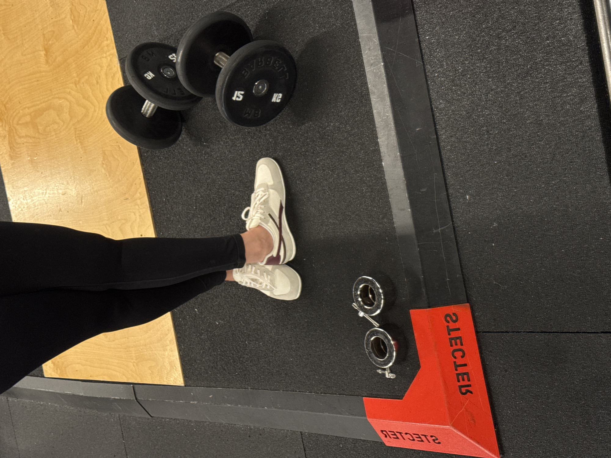 In an indoor fitness facility, possibly located in Sochi, Russia, a person stands on a black rubber mat. The person is wearing black leggings and white athletic shoes with burgundy accents. To the right of the person are two black dumbbells, each with weight indicators of "15" and "KG". In the foreground, near the bottom left of the image, are two chrome barbell collars. A red corner marker with white lettering that appears to be "RECKLESS" (when viewed in reverse) is also present. The background consists of a wooden platform and the edge of a black rubberized structure.