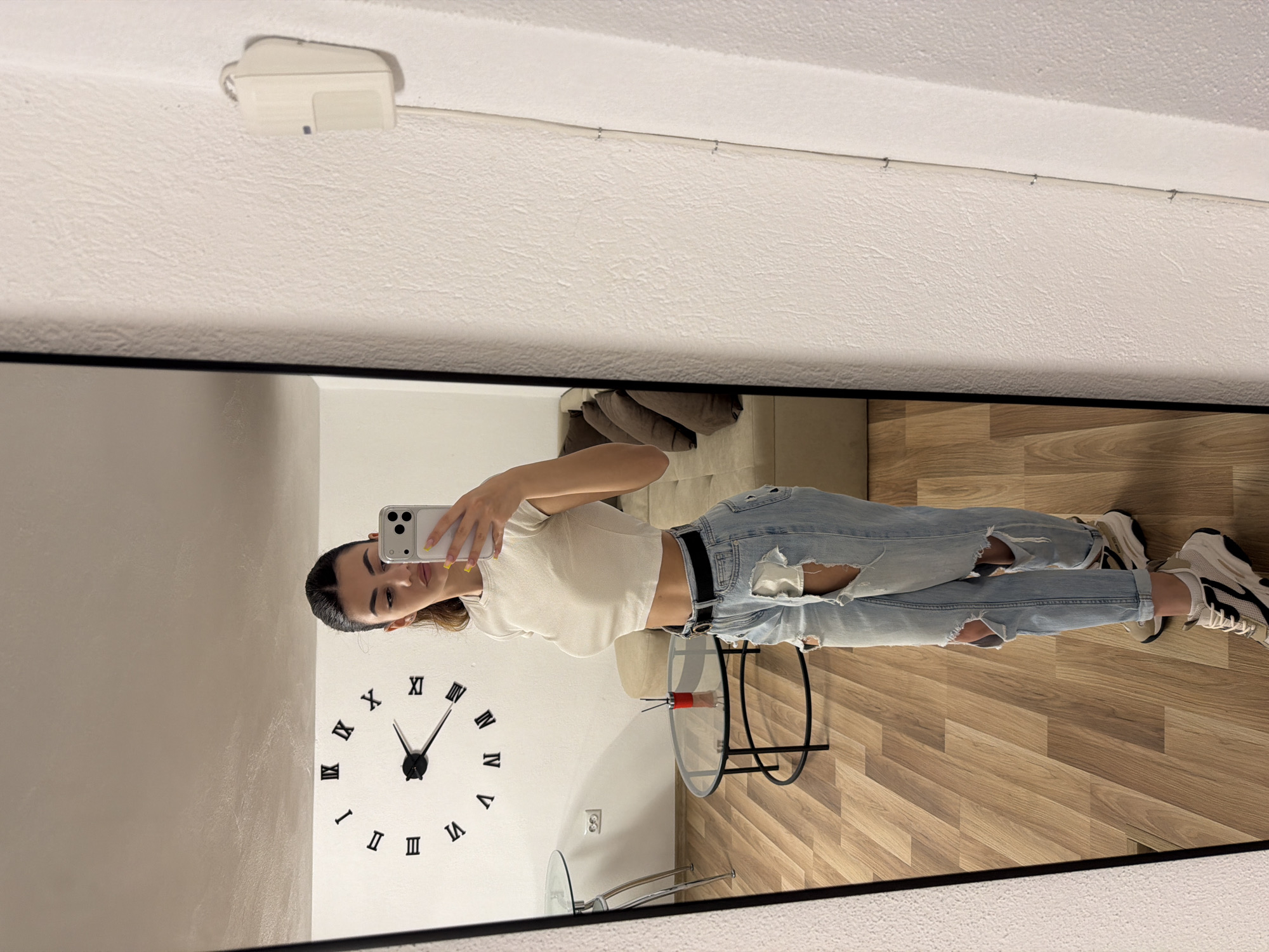 A young woman is taking a selfie in a mirror. The mirror shows a room with a clock on the wall. The woman is wearing a white crop top and ripped jeans with a black belt. She has sneakers on her feet. The room is not identifiable as being in the city of Gabrovo, Bulgaria. The mirror has a black frame. There is a light fixture on the ceiling above the mirror. The reflection shows furniture: a couch, a glass table and flooring with a wood grain pattern. The clock has black roman numerals. The woman is holding her phone up in her right hand.