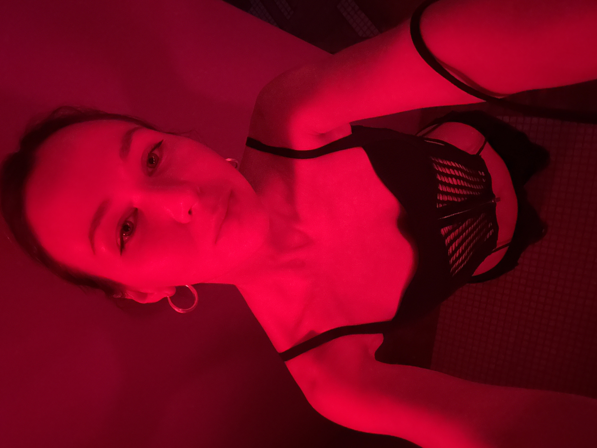 A female individual, appearing to be in her twenties or thirties, is depicted in a high-angle, eye-level self-portrait. She possesses dark hair, styled away from her face, revealing prominent eyebrows and visible eyeliner. Silver-toned hoop earrings are worn in both ears. The individual is dressed in black undergarments, including a bralette with thin shoulder straps. Layered over or integrated with this is a black structured garment, potentially a corset or bustier, featuring a ribbed or woven texture across the front. A thin black strap, likely from accompanying underwear, is visible below the primary torso garment. Her left arm is visible along her side, while her right arm extends towards the bottom right, appearing to hold the camera or phone. The entire scene is saturated in a monochromatic deep red light, originating from an unknown source. The background consists of a featureless wall, rendered crimson by the lighting. Subtle textural patterns, possibly from wallpaper or fabric, are discernible on the wall to the right and bottom right of the frame. The lighting casts a strong red hue across the subject's skin and the immediate surroundings. The specific indoor location is undetermined; it is in the city of Unknown, Unknown.