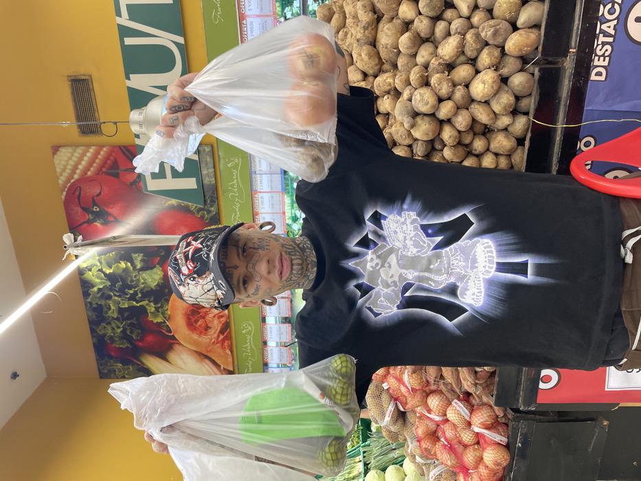 The image captures a person in what appears to be the produce section of a grocery store or supermarket. The image is oriented sideways, with the top of the scene to the left and the bottom to the right.
The central figure is a person with extensive facial and neck tattoos, covering areas around their eyes, nose, cheeks, chin, and neck with various patterns and possibly script. They are wearing a black short-sleeved t-shirt with a large, bright, and glowing graphic print on the front, depicting a stylized character or emblem with radiating light effects. They also wear a baseball cap with a complex, multi-colored design, and prominent ear gauges are visible in both ears. The person is looking directly at the camera with a neutral expression.
They are holding up two clear plastic bags, one in each hand, towards the viewer. The bag in their left hand (viewer's right) contains several light brown, rounded items, likely potatoes. The bag in their right hand (viewer's left) holds some green, spherical items, possibly limes or small green apples, and a bright green plastic bottle or container is also visible inside the bag.
In the background, to the right of the person, a large pile of loose, light brown potatoes is displayed in a bin. Behind this, a blue sign with white text is partially visible, showing the word "DESTACA" (Spanish for "highlight" or "featured"). A red, circular object is also partially visible on the far right.
To the left of the person, a yellow wall or ceiling section is visible at the top, featuring a rectangular air vent. Below this, large graphic posters decorate the wall, depicting various fruits and vegetables, including a prominent image of a red tomato with corn, green leafy lettuce, and a pumpkin or squash. White shelving units with price tags are visible in this area, displaying prices such as "3,999.00" and "1,299.00", along with Spanish text like "OFERTA" (offer) and "FERIA".
Below the person's hands, more produce displays are visible. Stacked mesh bags filled with brown, round produce (likely onions) are arranged on shelves, and below them, fresh green leafy vegetables are also displayed. The overall setting indicates a brightly lit indoor environment, characteristic of a supermarket.