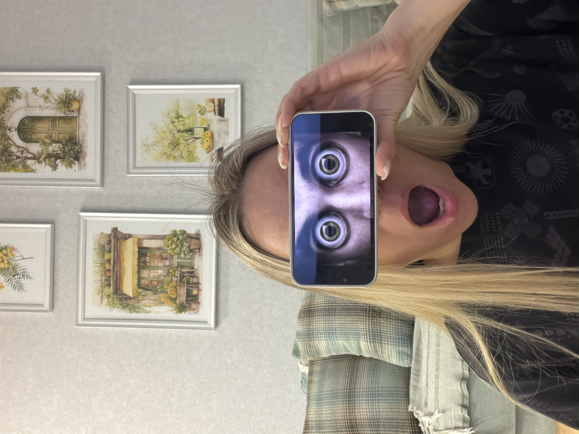 The image shows a person, identified as female with blonde hair, holding a smartphone in front of their face in an indoor setting in Votkinsk, Russia. The smartphone screen prominently displays a close-up of two eyes with widely dilated pupils, creating a surreal and exaggerated effect where the phone's eyes replace the person's own. The person's mouth is open wide, as if in a scream, gasp, or comedic expression. Her left hand, with white-painted fingernails, holds the phone steady. She is wearing a dark top with light, abstract patterns and a green, blue, and brown plaid shirt over it.
In the background, a light-colored, textured wall is adorned with four framed pictures. These appear to be watercolor prints or paintings depicting rustic, Mediterranean-style outdoor scenes, including: an arched green door surrounded by lush foliage, a still life with fruit and possibly a bottle on a table, a building with a yellow awning, and a floral arrangement featuring yellow blooms. The overall lighting suggests an indoor daytime environment. The entire image is rotated 90 degrees counter-clockwise from its natural upright orientation. No visible text is present.