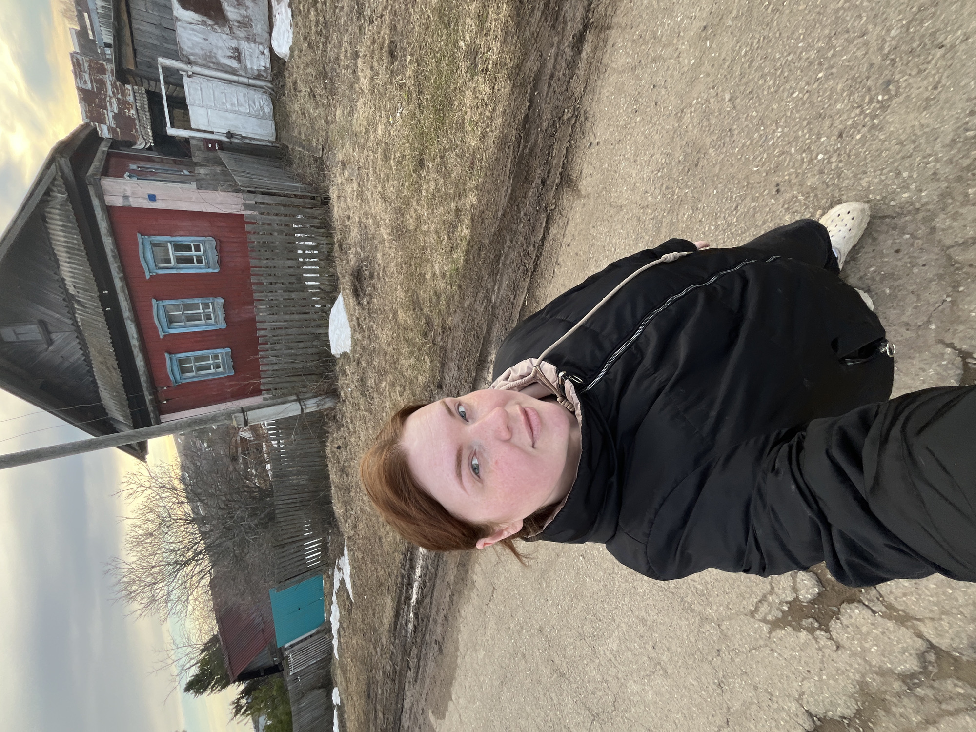 A young woman with reddish-brown hair and fair skin, wearing a black puffy jacket and white Crocs, stands on a cracked asphalt road. Behind her is a rural scene with weathered wooden buildings. A red building with blue-trimmed windows and a gray fence is visible to the left. Patches of snow are sparsely scattered in the surrounding dirt and dry grass. The sky is overcast. The location is Павловка, Russia.