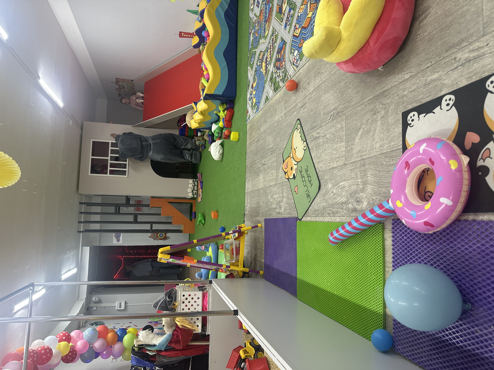The image displays a lively indoor children's play area, brightly lit by fluorescent lights on the white ceiling. The flooring is a patchwork of wood-patterned laminate, green artificial grass, and colorful textured mats in green and purple, alongside a black mat adorned with cartoon animal faces.
In the central part of the room, a person in a grey, fuzzy animal costume (resembling a mouse or bear) is seen from behind, leaning against a wall next to a window through which a child's face is partially visible. To their right, a large blue and yellow soft play structure, possibly a ball pit, is filled with colorful balls and sits on the green artificial grass. Further back, another child is visible near a red-painted wall section. A sizable rug depicting a detailed city map with roads and buildings covers a portion of the wood laminate floor, with an orange ball and a large yellow plush item resting on it.
In the foreground, a collection of toys includes a pink inflatable donut with colorful sprinkles, a striped blue and pink inflatable stick, a light blue balloon, and a small blue ball. A green mat clearly displays the text "WELCOME HOME" in white lettering. To the left, a string of multi-colored balloons (red, pink, yellow, blue, white) is arranged near a silver metal railing, beneath which various storage crates and other toys are visible. An orange and black staircase is present in the background on the right. The scene depicts a fun, activity-filled environment designed for children's play.