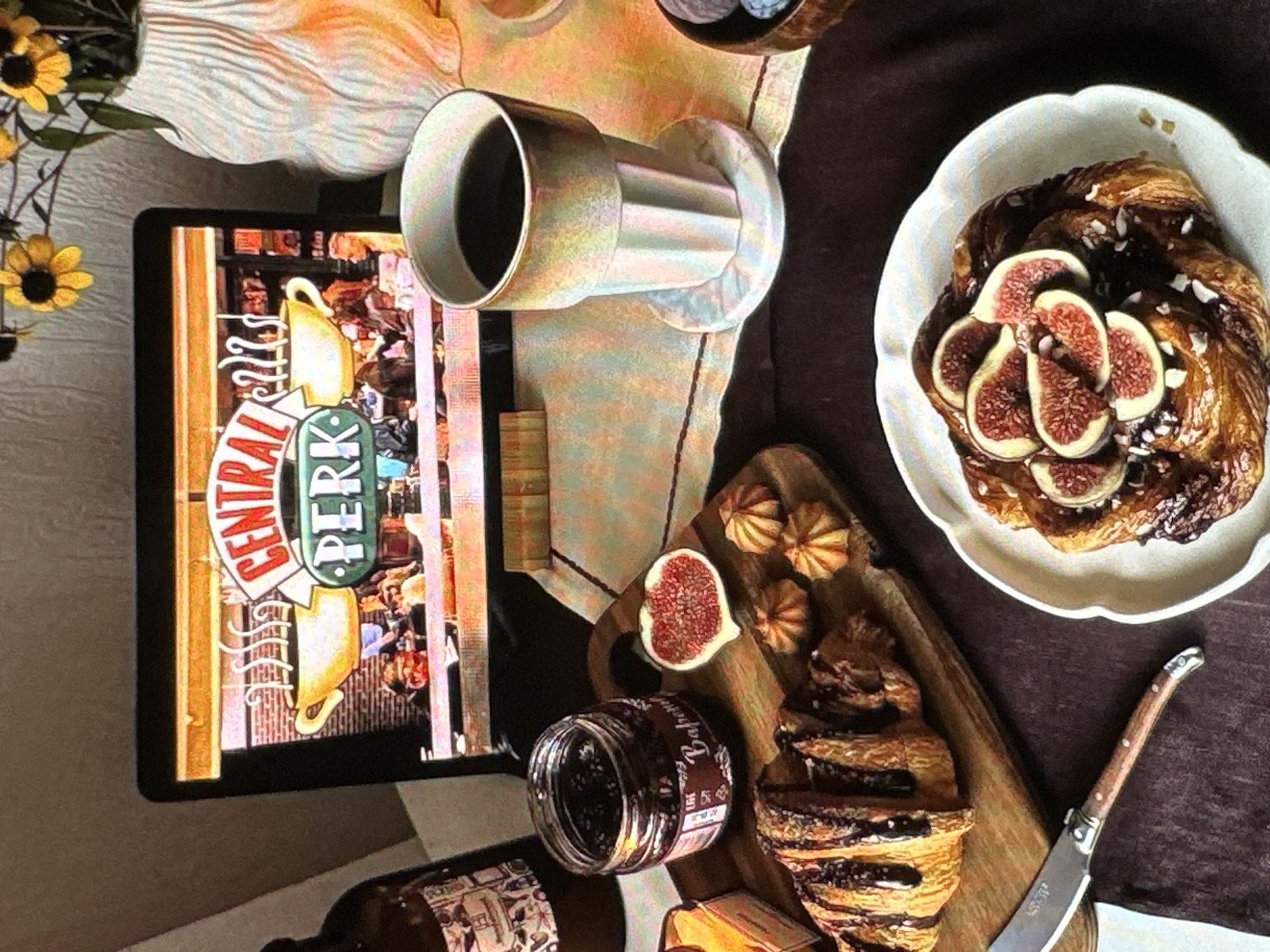 The scene presents a still life arrangement on a table, including food, drink, and media items. The foreground displays a white plate with a pastry adorned with fig slices and sprinkled with what appears to be coconut flakes. Beside the plate sits a wooden board with an additional pastry, a jar of dark jam, and a halved fig. A Laguiole-style knife rests near the board. A mug of dark liquid sits adjacent to the board. An electronic tablet displays an image of the "Central Perk" logo from the TV show "Friends". There is a bottle of liquid, possibly coffee concentrate, with labeling written in a European language. Additionally, there is a small arrangement of sunflowers. The setting appears to be a home interior. The location, Yerevan, Armenia, is unknown.