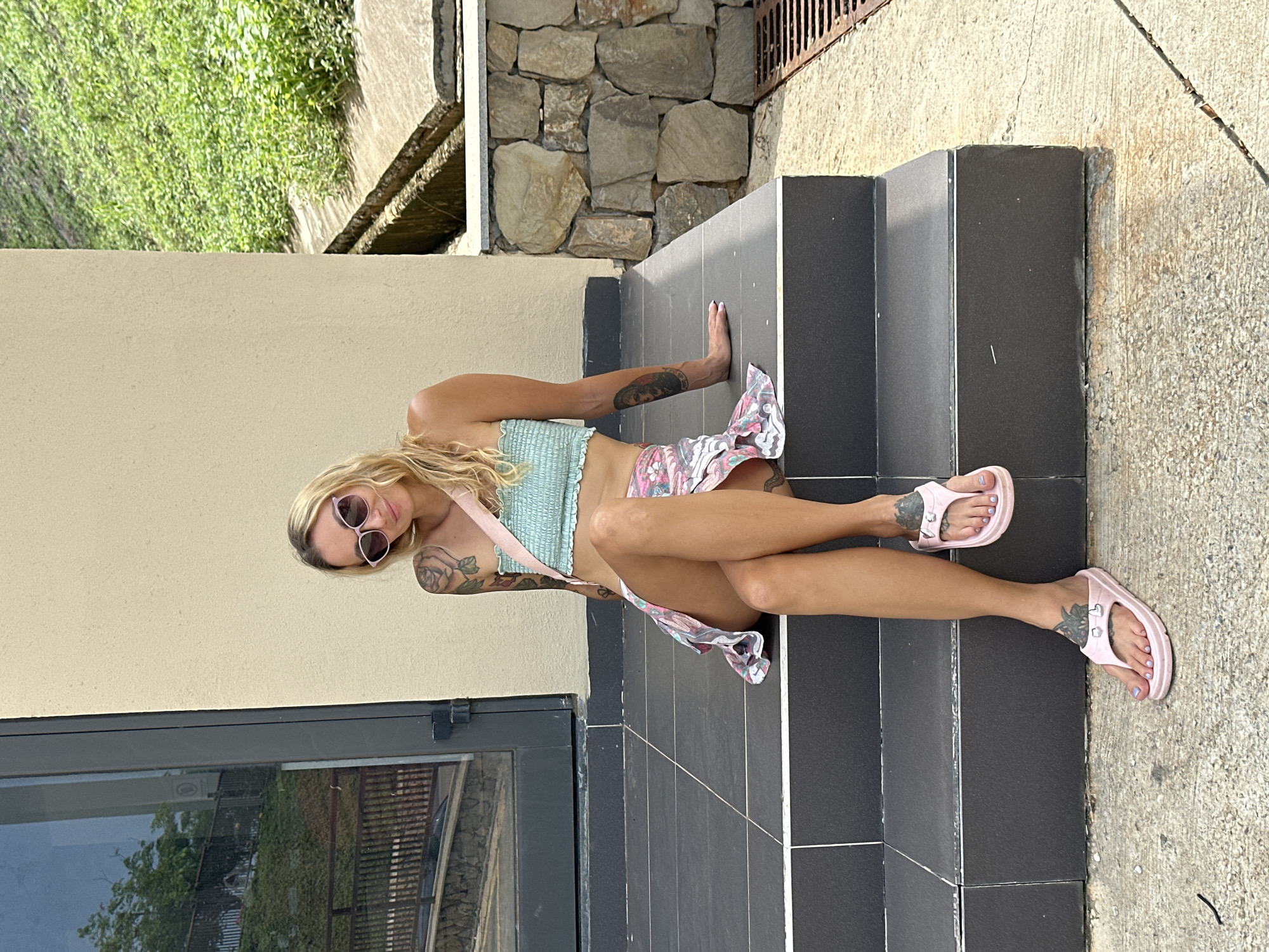 The image displays a woman seated on a set of dark grey tiled steps in Tien Thanh Ward, Vietnam. The woman has long, wavy blonde hair and a tanned complexion. She wears pink cat-eye sunglasses with dark lenses, which obscure her eyes, but a slight smile is visible. Her attire consists of a light mint green smocked bandeau crop top and a short, floral-patterned skirt primarily featuring pink, white, and green hues. A light pink strap, likely from a bag, crosses her chest.
Visible tattoos include a large rose design on her right upper arm/shoulder and a portrait or figure on her left forearm, along with other smaller designs. She also has small tattoos on the tops of her feet. She is wearing pink platform flip-flops, and her toenails are painted with light purple polish.
The woman is positioned on the third step from the bottom, with her right leg slightly extended forward and her left leg bent, with her foot resting near her right ankle. Her left hand rests on the step behind her, while her right arm is casually placed across her lap.
To the left of the woman is a building with a light beige/cream wall and a dark-framed, reflective glass door or window, reflecting trees and sky. To her right, a lush green hillside covered with dense vegetation is visible, alongside a concrete drainage ditch and a low wall constructed from irregularly stacked, light-colored stones. A metal grate is present on the concrete ground level. The steps are dark grey with light-colored grout lines and lead down to a textured concrete sidewalk or path.