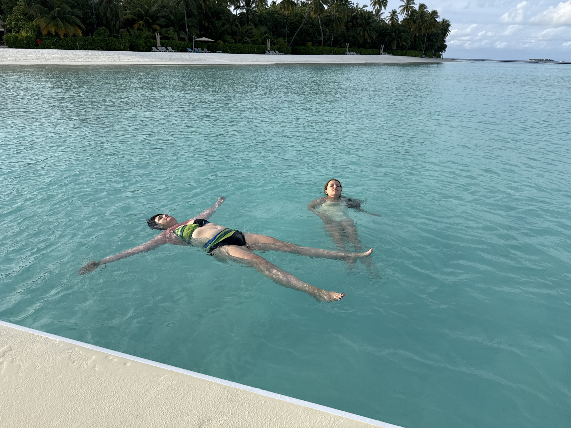 Two women are floating on their backs in clear turquoise water. The woman in the foreground is wearing a green and black striped bikini and has her arms and legs spread wide. The woman in the background is wearing a light-colored swimsuit and her face is tilted upwards towards the sky. In the background, a white sandy beach is lined with palm trees and lounge chairs, with a calm ocean stretching to the horizon under a cloudy sky. The scene appears to be the shore of the Maldives.