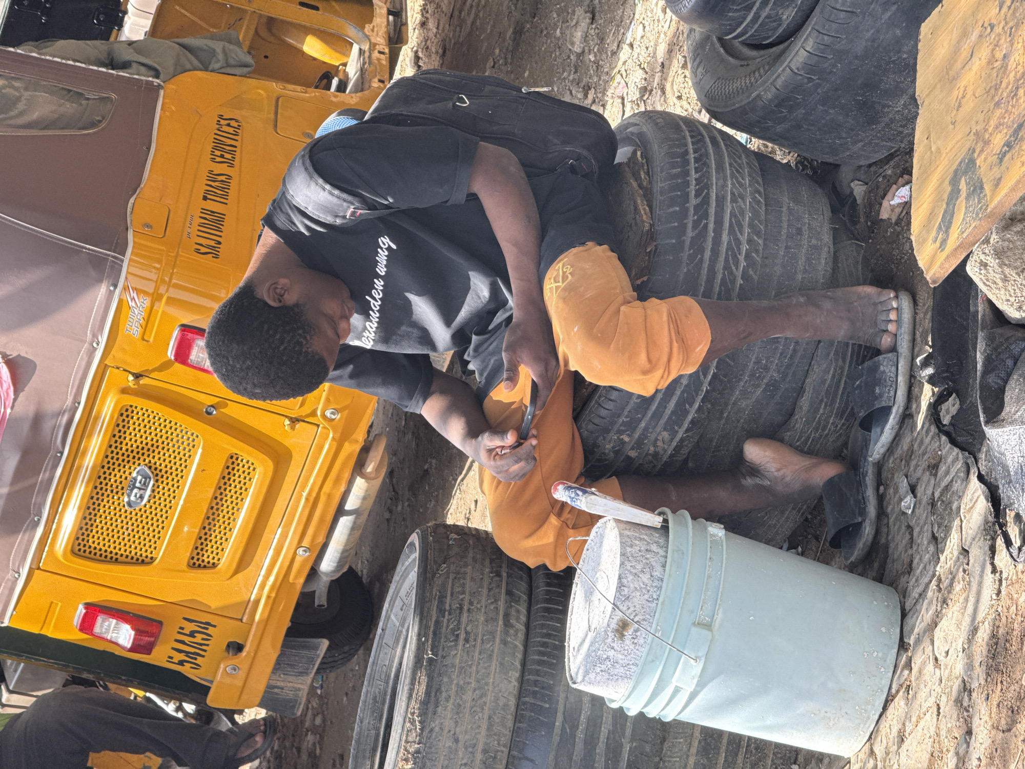 A person, wearing an orange garment and a black t-shirt with white text, sits on a pile of tires. The t-shirt has a logo on the left breast and white lettering that appears to read "Alexander Mwng". The person is holding a tool, possibly a knife, and working on something in their lap, which is obscured by their body. To the right of the person is a light blue bucket with a white lid, and a metal rod sticking out of it. A yellow vehicle, possibly an auto-rickshaw, is visible to the left. The side of the vehicle has black text that reads "SAJJIDAH TRANS SERVICES" and "PETROL". Below this, there's a smaller logo with the word "TWIN" and "SPARK". The rear of the vehicle has a license plate with the number "54454". The scene appears to be outdoors, with dirt and debris on the ground, suggesting a rural or informal setting. The lighting suggests it could be daytime, with no specific indicators of time of day or weather. The overall impression is one of manual labor or repair work in an industrial or utilitarian environment.