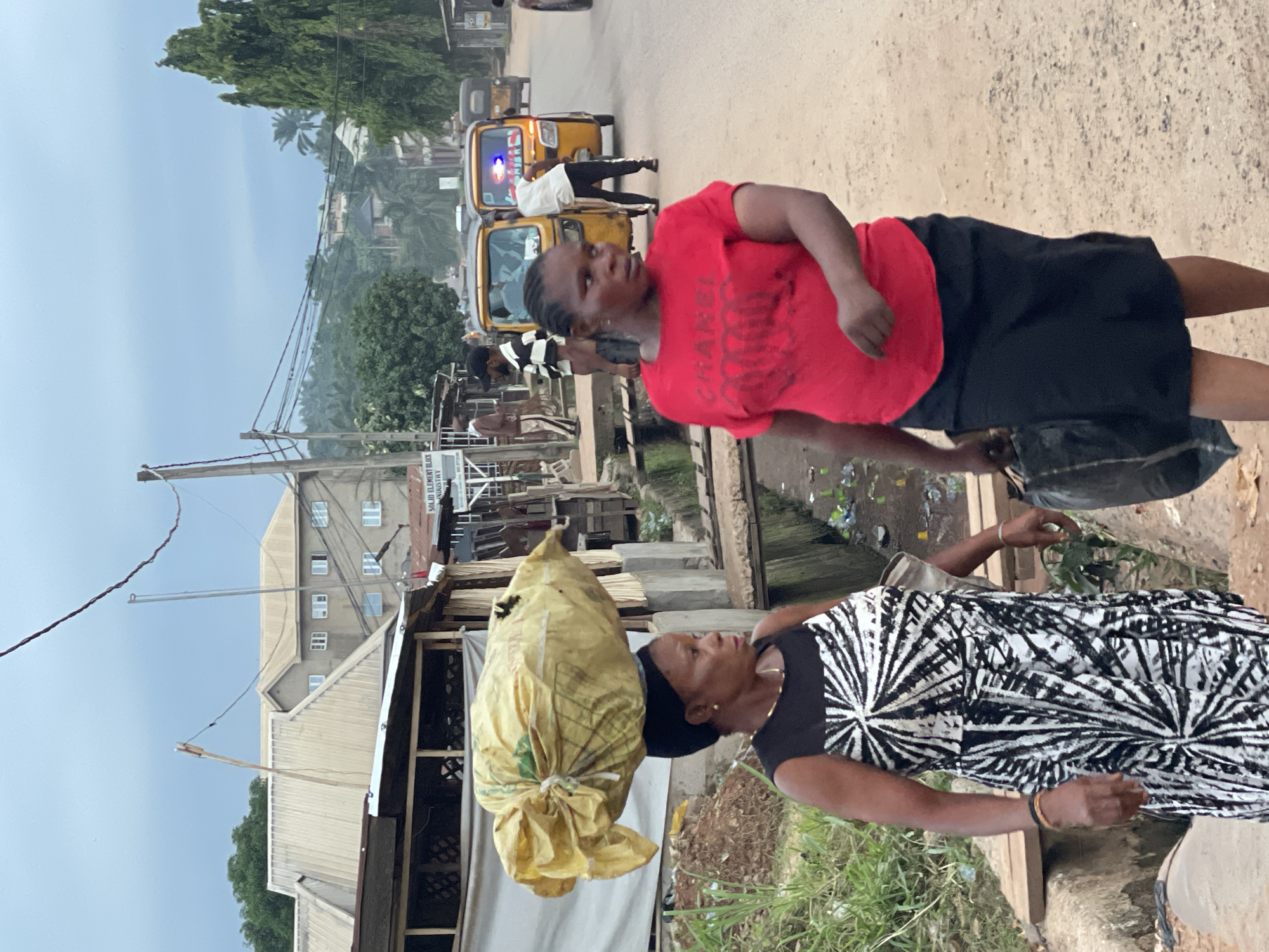 In Nkpor, Nigeria, a woman in a black and white patterned dress carries a large yellow sack on her head. Another woman, wearing a red t-shirt and black skirt, walks beside her carrying a black bag. A yellow auto rickshaw taxi passes in the background. Shops and residential buildings are visible. The sky is overcast.