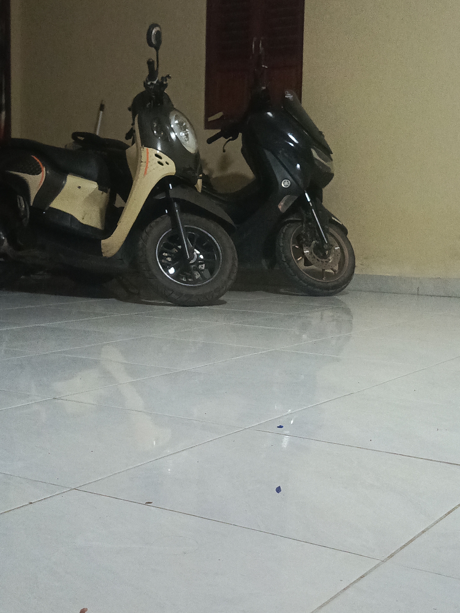 Two motorcycles are parked next to each other in front of a light-colored wall. The motorcycle on the left is a beige and black scooter with a round headlight. The motorcycle on the right is a black scooter with a more angular design. Both motorcycles have their wheels on a light-colored tiled floor. The scene appears to be an indoor or covered area, possibly a garage or parking space. The lighting is dim, creating shadows on the floor. The location is identified as Merubung, Indonesia.
