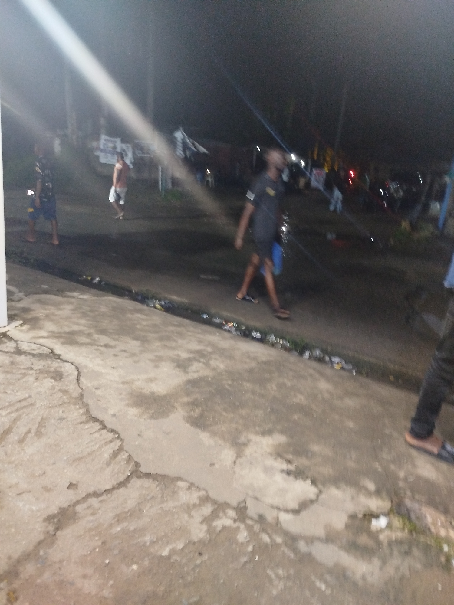 The image depicts a nighttime scene in Ikot Mbonde, Nigeria. The location appears to be a street with a concrete sidewalk in the foreground. Several people are present, with some walking along the street. One individual is wearing dark clothing, while another is wearing lighter-colored shorts. The street is dimly lit by what appears to be a streetlight. There is some trash on the ground along the side of the street. In the background, there are indistinct structures.
