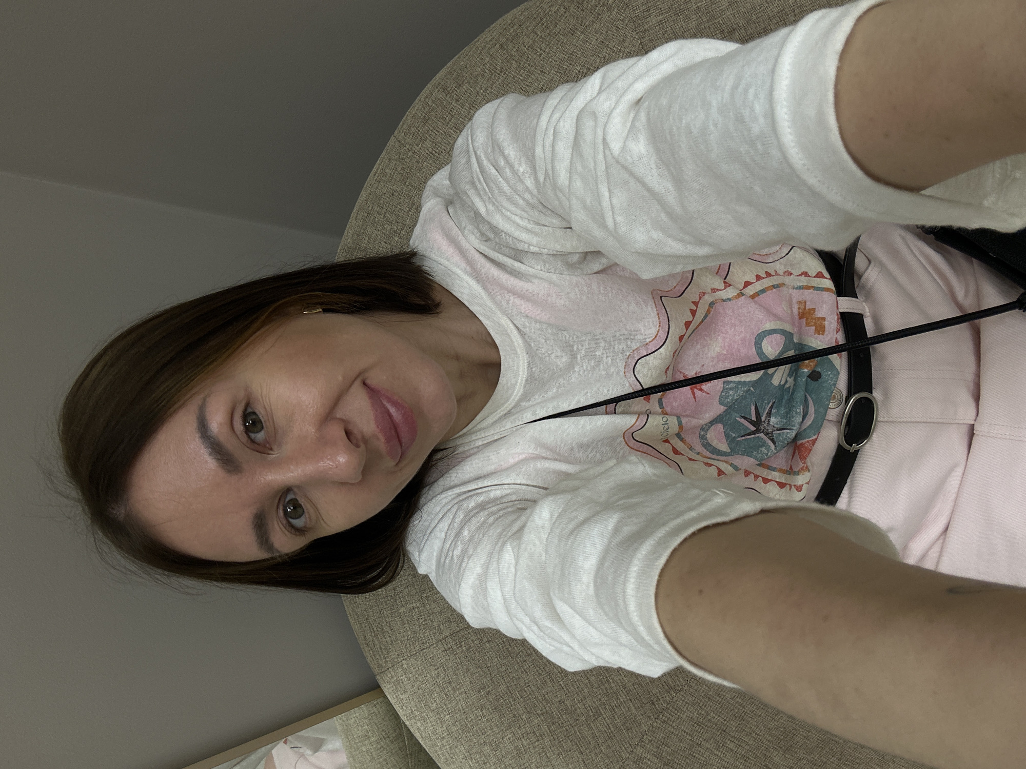 A woman with shoulder-length brown hair and light skin is captured in a selfie, seated in a beige armchair. She is wearing a white long-sleeved shirt with a graphic print featuring a cartoon-like animal and abstract shapes in pink, blue, and yellow. A black belt with a silver buckle cinches her waist, and she has light pink pants on. A black strap is visible, possibly from a bag. The background appears to be an indoor setting, with a grey wall and a corner of a mirror reflecting a portion of the scene. The lighting is soft and diffused. The image does not contain any specific landmarks or clues that would allow for identification of a location within the city of Izhevsk, Russia.