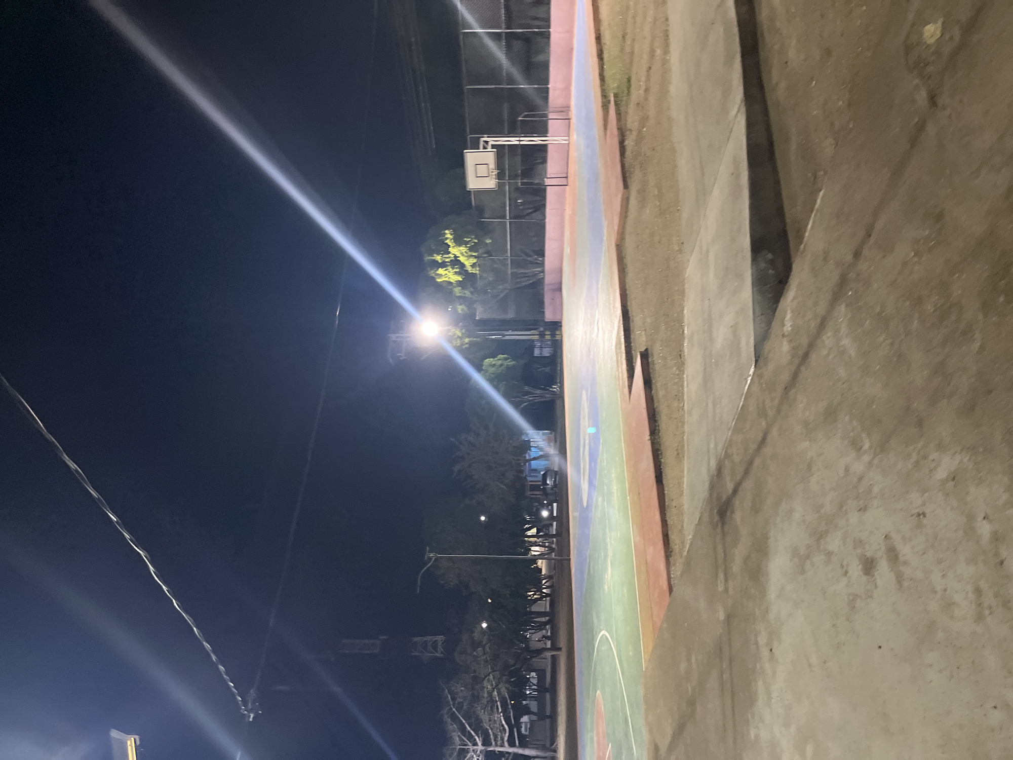 This image depicts an outdoor basketball court at night in Santa Ana, Venezuela. The court is illuminated by artificial lights, casting long shadows and creating bright streaks across the dark sky. The court surface is multi-colored, with sections of green, blue, and light purple. A basketball hoop and backboard are visible on one end of the court, with a chain-link fence behind it. Trees line the perimeter of the court, and a transmission tower can be seen in the background. In the foreground, there are concrete pathways and benches. The scene appears deserted.