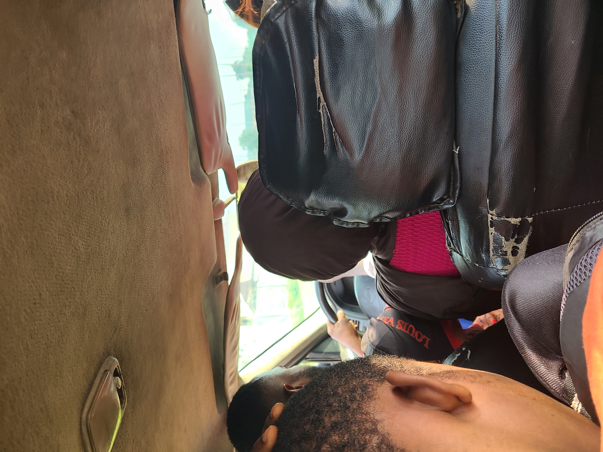 The image depicts the interior of a vehicle in Karu, Nigeria, with passengers seated inside. A person wearing a dark hijab and a pink top is in the driver's seat, hands on the steering wheel. To the left, two individuals are visible from behind, facing forward. One of them has a black t-shirt with white and red lettering that appears to say "LOUIS VUITTON". The vehicle's interior shows signs of wear, with damaged leather seats and a worn headliner. The overall lighting suggests it is daytime, with natural light entering through the windows. The background visible through the windshield consists of greenery and structures, indicative of an outdoor, possibly urban or semi-urban, environment.