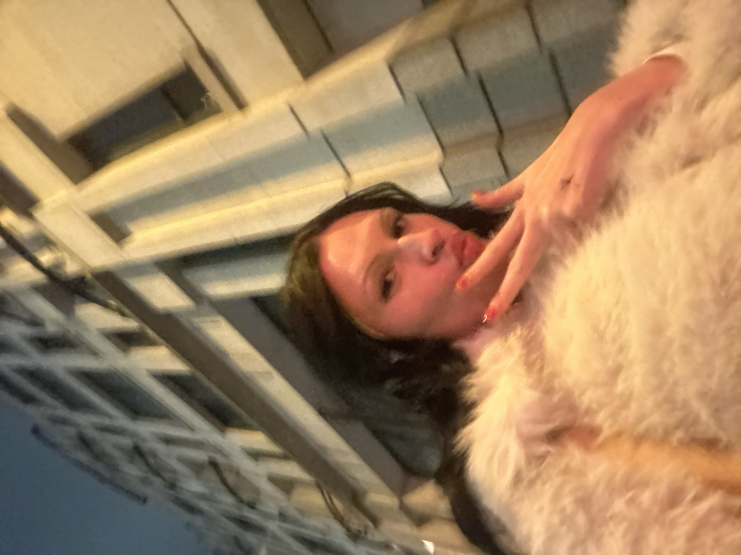 A low-angle, tilted shot shows a woman with long dark hair, wearing a fluffy white garment, possibly a coat or sweater. Her hand is near her face. The background features a building with a grid-like architectural pattern, suggesting an urban environment. The lighting is warm, with shadows emphasizing the building's structure. The woman's pose and the upward angle create a casual, selfie-style image.