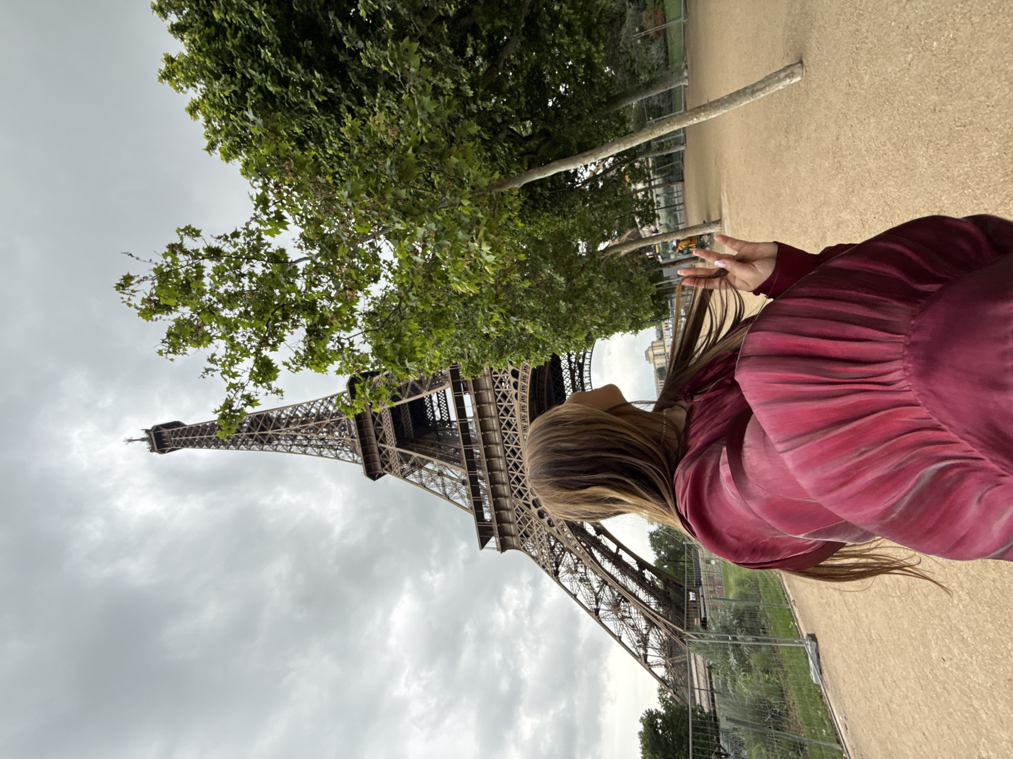 A person poses with the Eiffel Tower in Paris, France. The tower is visible behind the person. The sky is cloudy. Trees are visible around the base of the tower. The person is wearing a maroon shirt and is making a peace sign with their hand. They have shoulder-length brown hair. The ground appears to be sand.