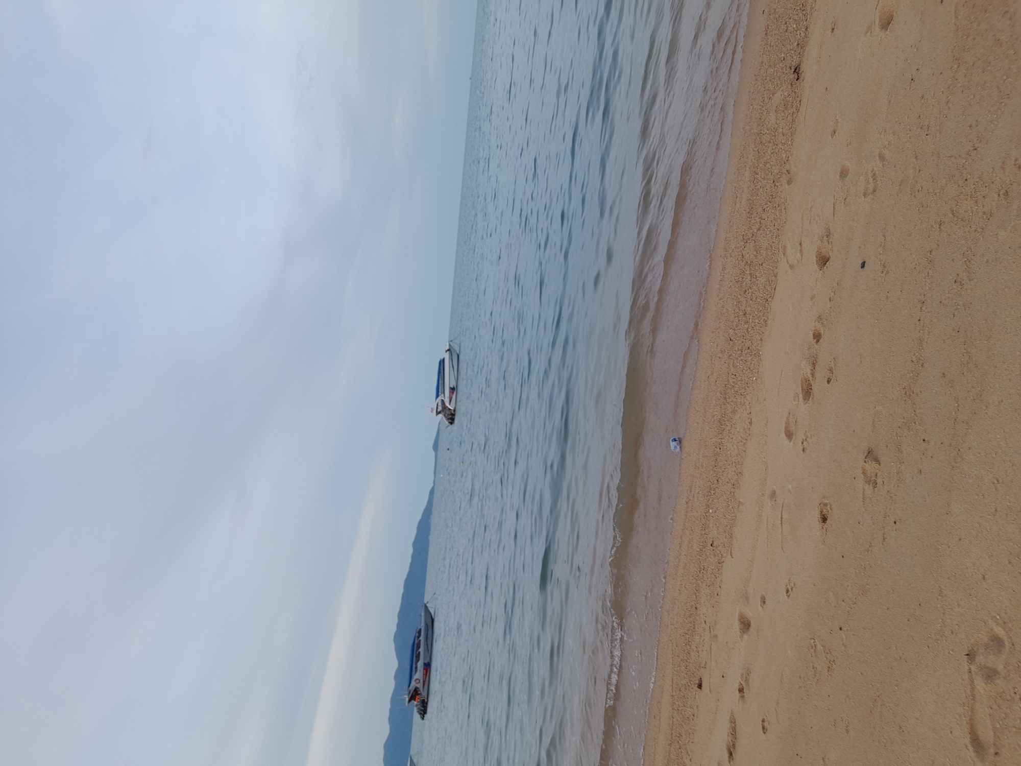 This is an image of a tropical beach scene in Baan Tai, Thailand. The image is oriented vertically, with the ocean on the left and the sandy beach on the right. There are two small speedboats in the water, one closer to the shore than the other. The water is a calm blue, with gentle waves lapping at the sand. The beach is made of light-colored sand, and there are footprints visible on the sand, suggesting recent human activity. In the distance, a hazy outline of mountains can be seen on the left side of the frame, characteristic of the island landscape. The sky is overcast with soft, diffused light, indicating either early morning or late afternoon, or a cloudy day. There is no visible text in the image. The overall atmosphere is tranquil and serene.