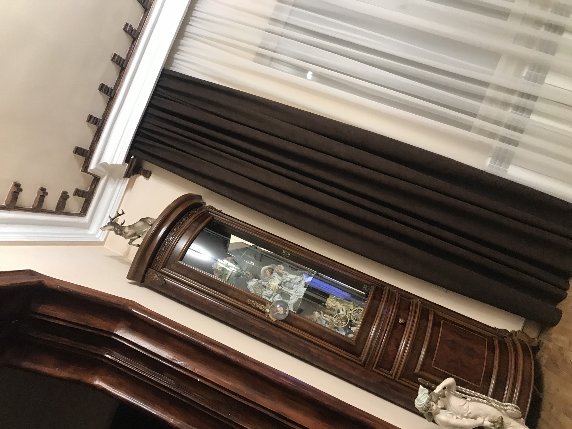 The image shows an interior scene with a curio cabinet against a beige wall, situated below a window with white sheer and brown blackout curtains. The cabinet is dark wood and houses figurines and decorative objects inside. Atop the cabinet is a deer statue. A dark wood molding is visible in the foreground. On the floor next to the cabinet is a statue of a reclining figure. The image suggests a well-appointed interior space, and the style of the cabinet and décor suggests a traditional or classic aesthetic. The location is not identifiable from the image.