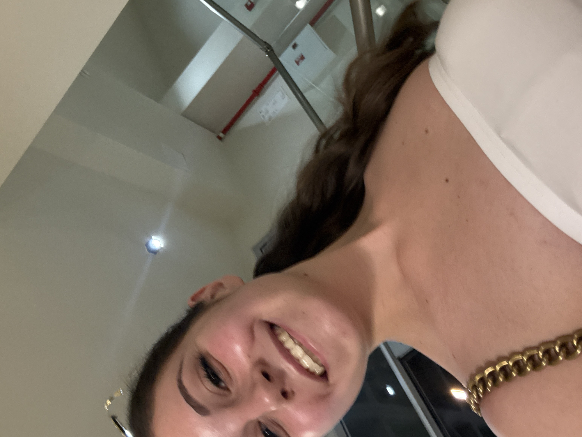 The image is a close-up, selfie-style shot of a young woman with brown hair pulled back in a ponytail. She is smiling, showing her teeth which have dental aligners. She is wearing a white off-the-shoulder top and a gold chain necklace is visible on her left shoulder. The background appears to be indoors, possibly a modern building with metallic railings and cream-colored walls. There is a red pipe running along the wall. The lighting suggests an interior setting, possibly at night. There is no visible indication of the image being taken in Burgas, Bulgaria.