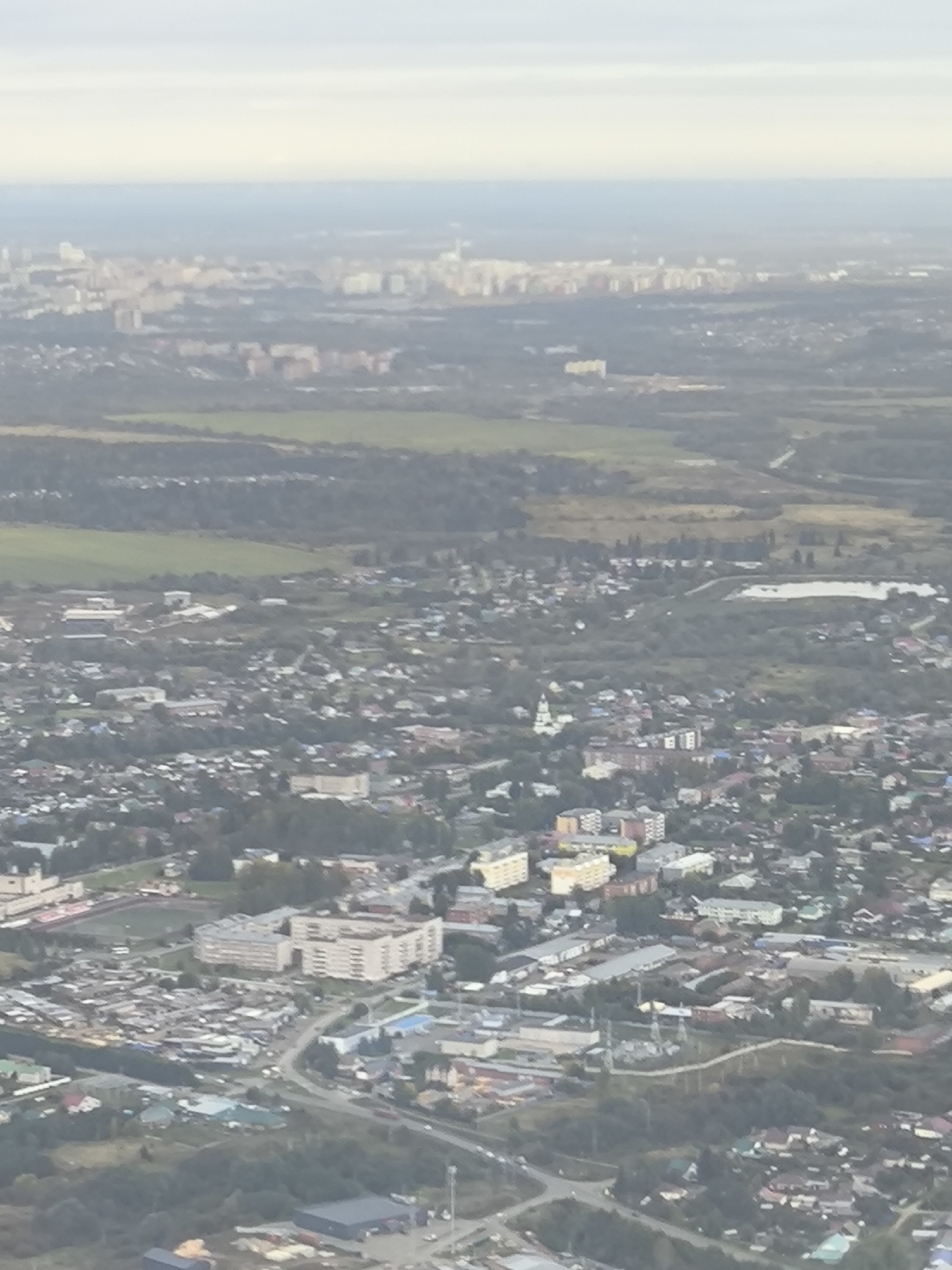 This is an aerial, high-angle, slightly blurry, daytime photograph of a sprawling urban landscape under a hazy sky. The foreground is dominated by a densely packed residential area with a mix of low-rise houses and several multi-story apartment buildings. Roads and smaller streets weave through the buildings, and there are areas of greenery interspersed throughout the town. Further into the midground, more apartment complexes and commercial or industrial buildings are visible, some with flat roofs and a uniform architectural style. In the background, the city extends further, with a cluster of taller buildings suggesting a more developed urban center or the outskirts of a larger city. Fields and patches of trees are visible between the developed areas, indicating a transition between urban and rural environments. The overall impression is of a moderately sized city or a large town with a blend of residential, commercial, and industrial zones. The lighting suggests it could be late afternoon or early morning due to the softness of the light and the hazy atmosphere.