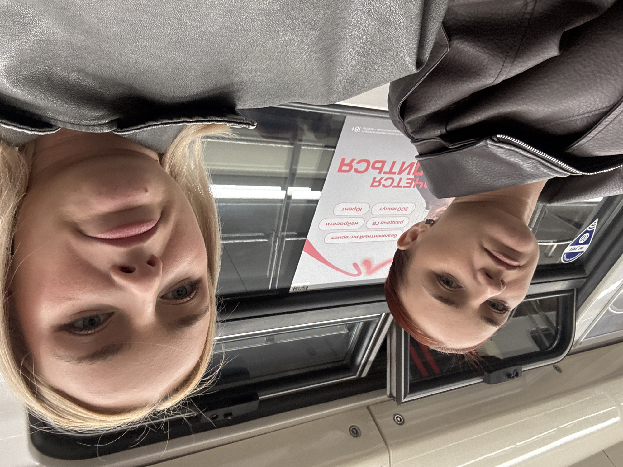 Two young women are seated next to each other, possibly on public transportation, based on the overhead lighting and window framing. The woman on the left has auburn hair and is wearing a dark leather jacket. She is looking directly at the camera with a neutral expression. The woman on the right has blonde hair and is also wearing a dark leather jacket. She is also looking towards the camera, with a slightly distressed or pouting expression. In the background, a sign with text in Russian is visible, indicating some sort of service or advertisement. The overall environment appears to be indoors, with artificial lighting.