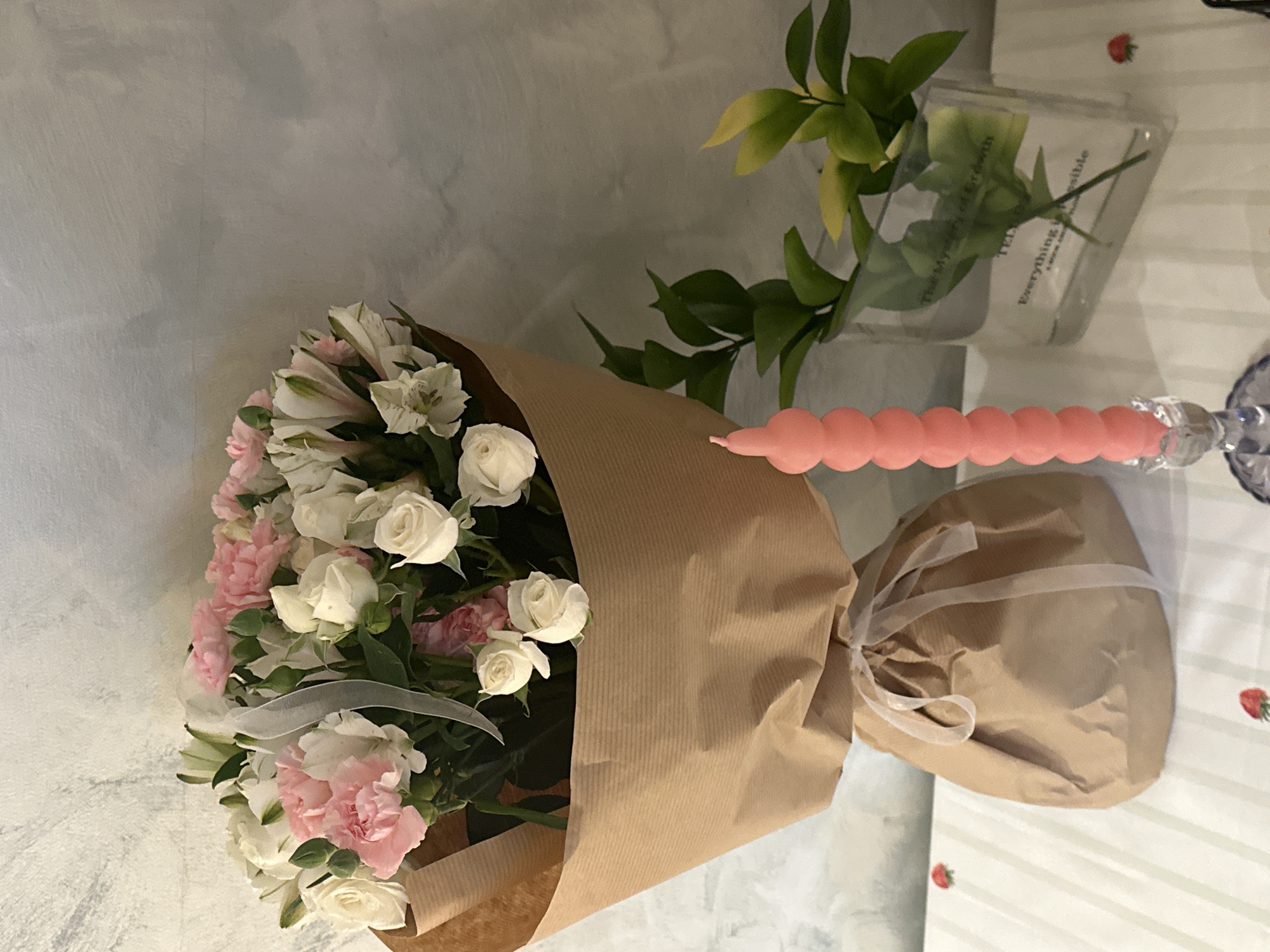 The image depicts a bouquet of white and pink flowers wrapped in brown paper, resting on a table. Next to the bouquet, there is a clear vase with green leaves and a decorative pink candle with a beaded design. The table has a light-colored tablecloth with strawberry patterns. The environment suggests an indoor setting, possibly a home or event space. The location of Šušanj, Montenegro, is not evident from the visual content of the image.