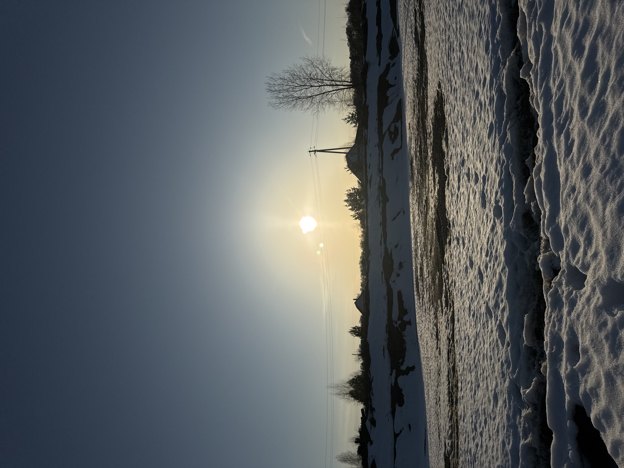 The image depicts a winter scene near Chaikovsky, Russia. A snow-covered field occupies the foreground, its surface marked with undulating patterns and occasional dark patches indicating exposed ground. The sun, low in the sky, shines brightly, its glare causing a halo effect. In the midground, a line of trees, a few buildings, and a utility pole are visible. The sky is a gradient of blue, lighter near the horizon where the sun is located. The picture is oriented vertically.