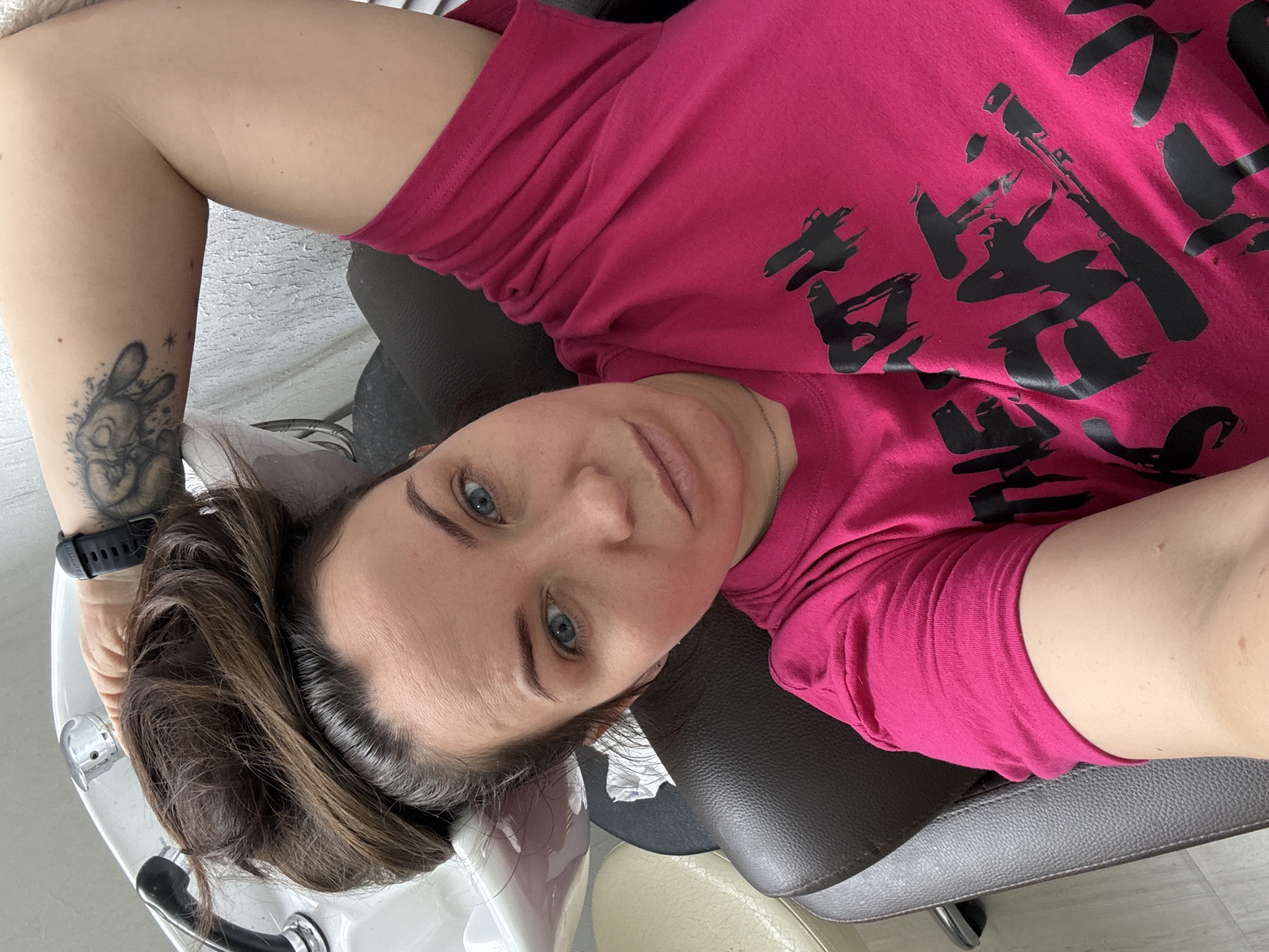 A woman with dark brown hair and blue eyes lies on her back in what appears to be a hair salon in Chaikovsky, Russia. She is wearing a bright pink t-shirt adorned with large, stylized black characters or symbols on the front. Her head is positioned over a white ceramic hair wash basin, suggesting she is about to have her hair washed or is receiving a hair treatment.
She looks directly at the camera with a neutral expression. Her left arm is visible, extended upwards, likely holding the camera to take a selfie. On her left forearm, there is a prominent tattoo of a cartoon-like bunny with stars. A black smartwatch, with "Huawei" discernible on its strap, is worn on her left wrist. She is resting on a dark brown, padded salon chair, with parts of a light-colored chair and the salon floor visible in the background. The lighting is even, indicating an indoor setting.