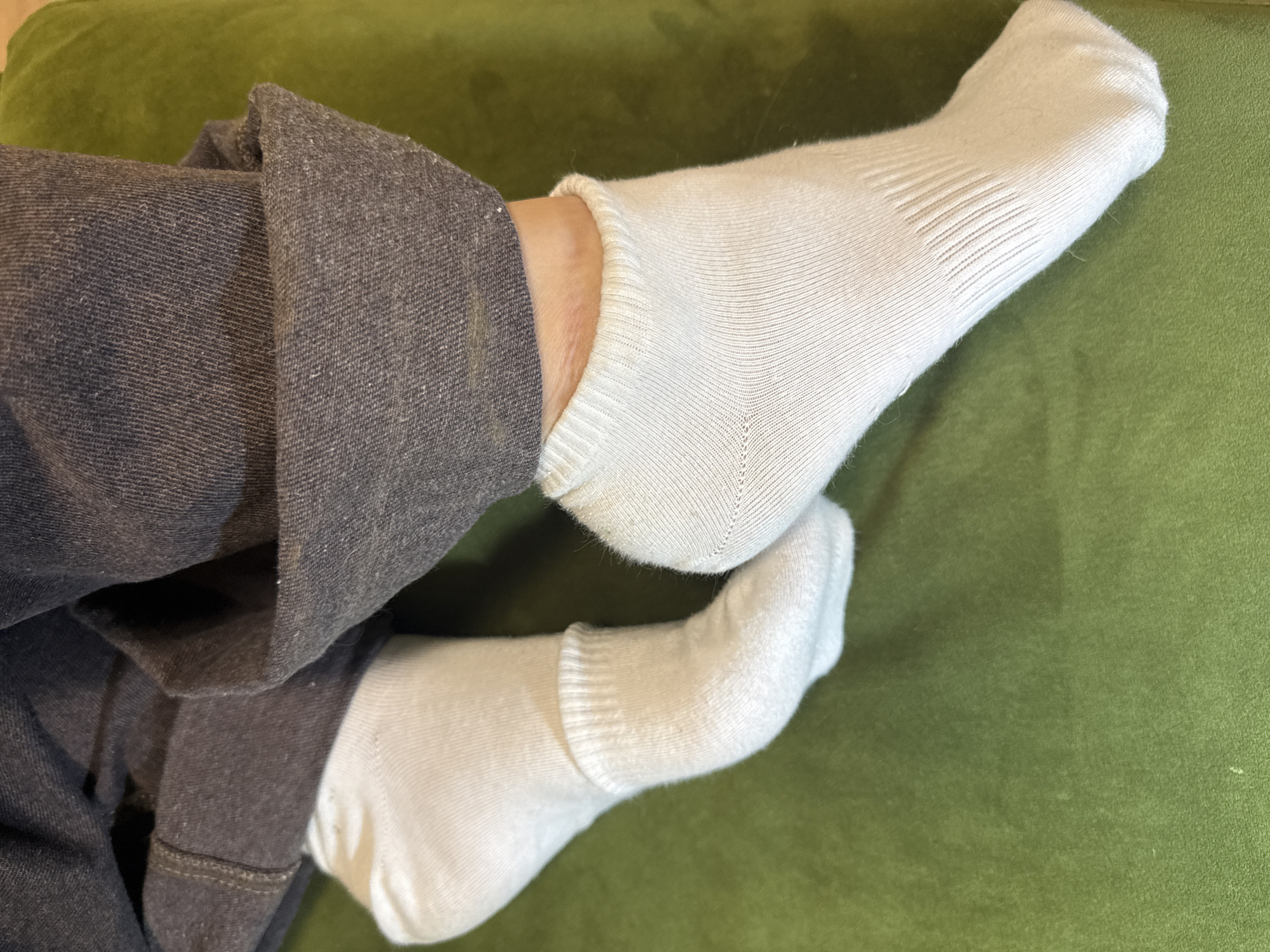 A close-up shot shows two legs wearing white socks and gray pants resting on a green surface. The composition focuses on the lower extremities of a person, with the texture and color of the fabrics being prominent. The background is a solid green, possibly a cushion or a piece of furniture. The location is not discernible from the provided image.