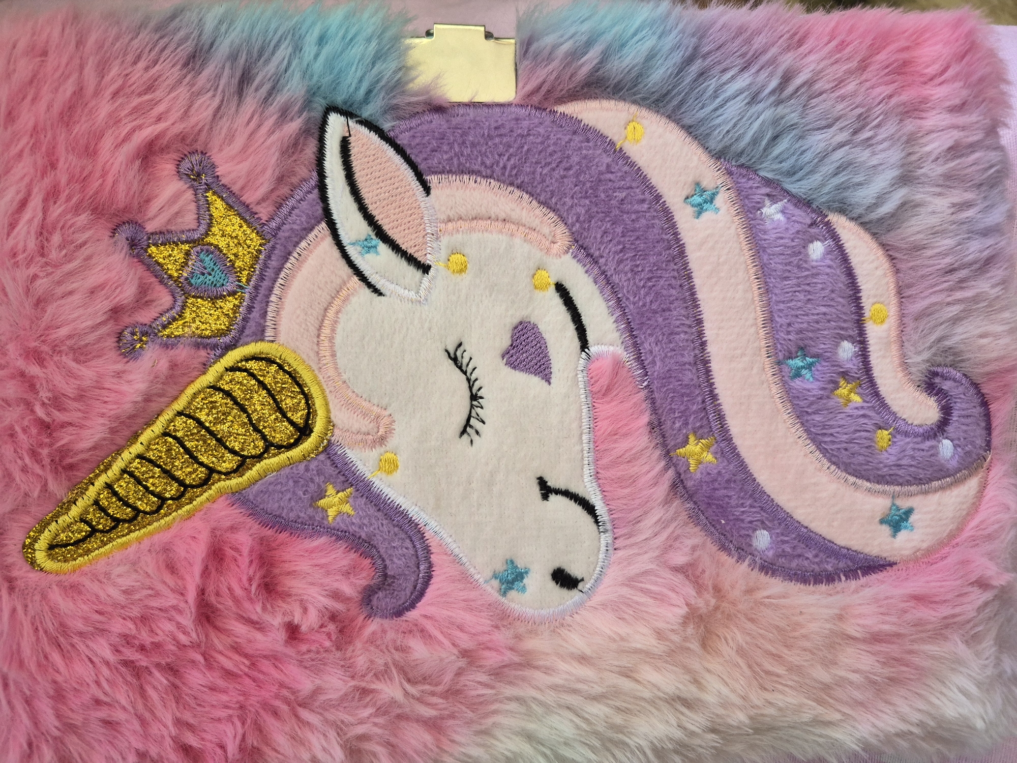 The image shows a close-up of a plush, rectangular object, possibly a notebook or diary cover. The background is made of long, soft fibers in shades of pink, white, and light blue. An embroidered unicorn design is centrally located. The unicorn has a white head with a closed eye and a heart-shaped marking. It has a gold glittery horn and crown. The unicorn's mane is striped in purple and pink, adorned with star designs in yellow and blue. A small gold clasp is visible at the top edge of the frame. The image provides no information about Tobolsk, Russia.