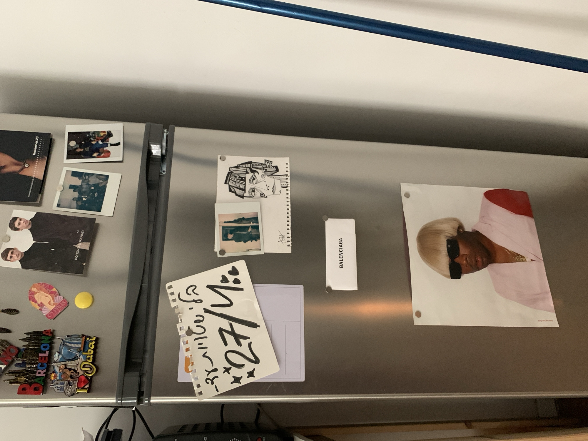 This image shows a silver refrigerator door covered with various items. These include photographs, drawings, brand labels, tourist souvenir magnets, and a handwritten note on a notepad. A poster of a person wearing sunglasses and a blond wig is attached to the upper right side of the refrigerator door. The environment appears to be a kitchen or dining area. There is no indication of any location in the city of Tel-Aviv, Israel.