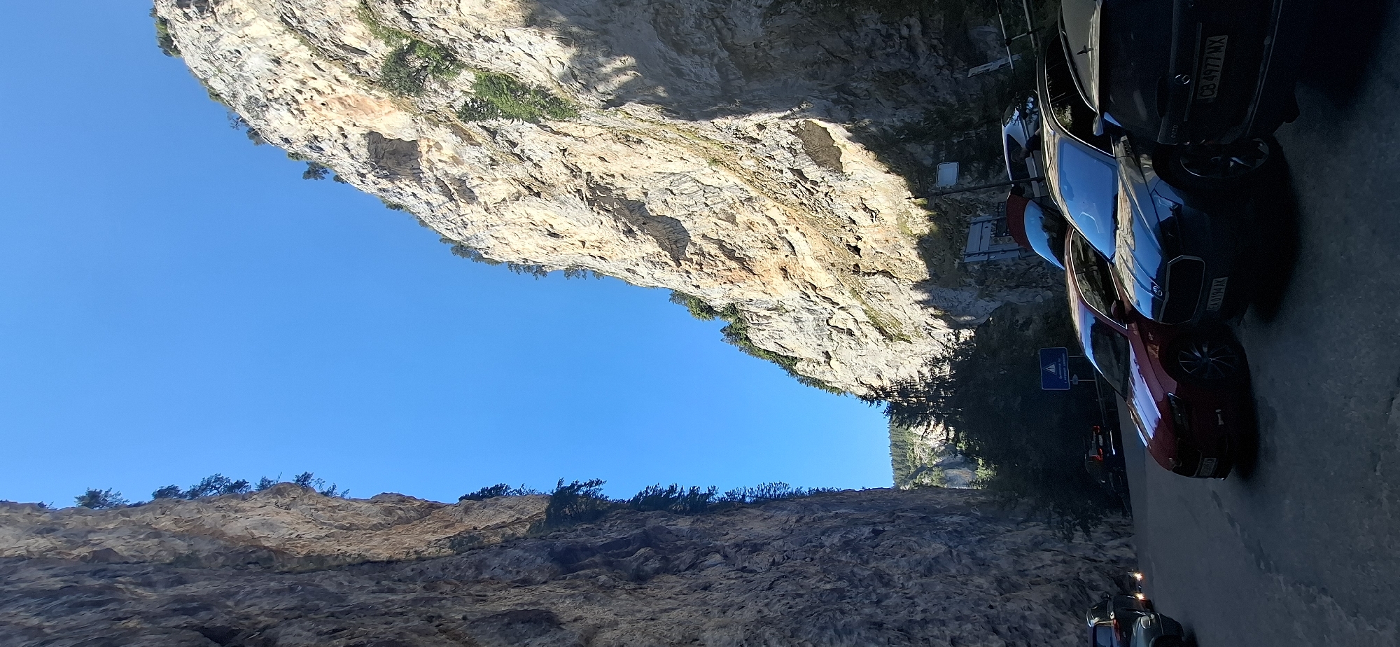 The image depicts a road winding through a narrow gorge with towering, rocky cliffs on either side. The sky above is a clear, bright blue. The cliffs are steep and irregular, with sparse vegetation clinging to their surfaces. In the foreground, several cars are parked on the side of the road. The cars are of various colors, including red and dark gray. One of the cars has a license plate that reads "EA 0194 AK". Another license plate is partially visible and reads "CB 4977 MX". A blue triangular road sign with a white symbol is visible on the left side of the road. The scene appears to be a scenic route or a point of interest, likely located in the city of Trigrad, Bulgaria, known for its dramatic gorges and natural landscapes. The overall impression is one of natural grandeur and a popular tourist destination.