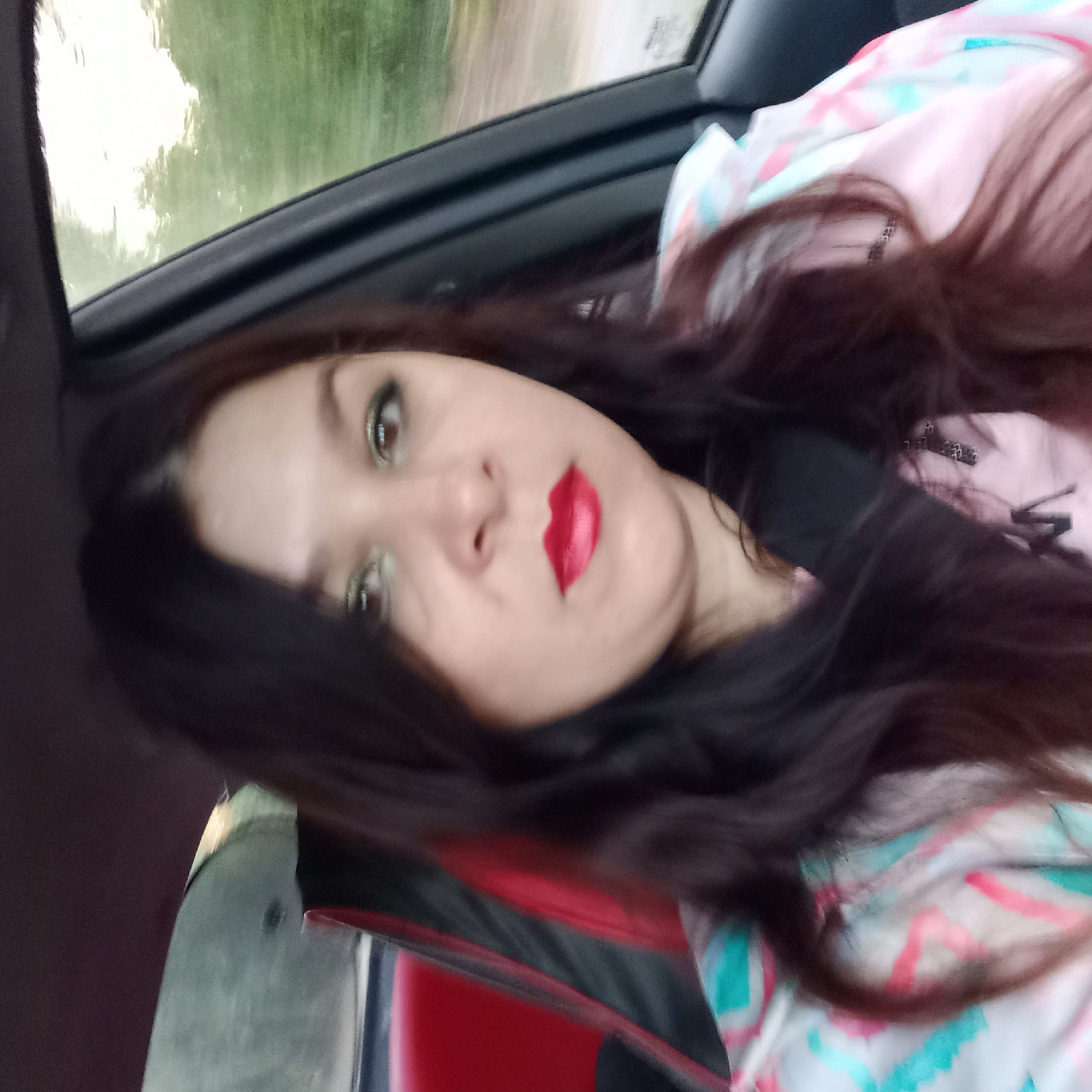 The image is a selfie taken from inside a car, likely in Kropyvnytskyi, Ukraine. A woman with dark hair and makeup, including green eyeshadow and red lipstick, is visible. She is wearing a pink and light blue patterned jacket over a pink shirt. The background shows trees and sky through the car window, suggesting an outdoor setting. The interior of the car is partially visible, with a red and black seat.