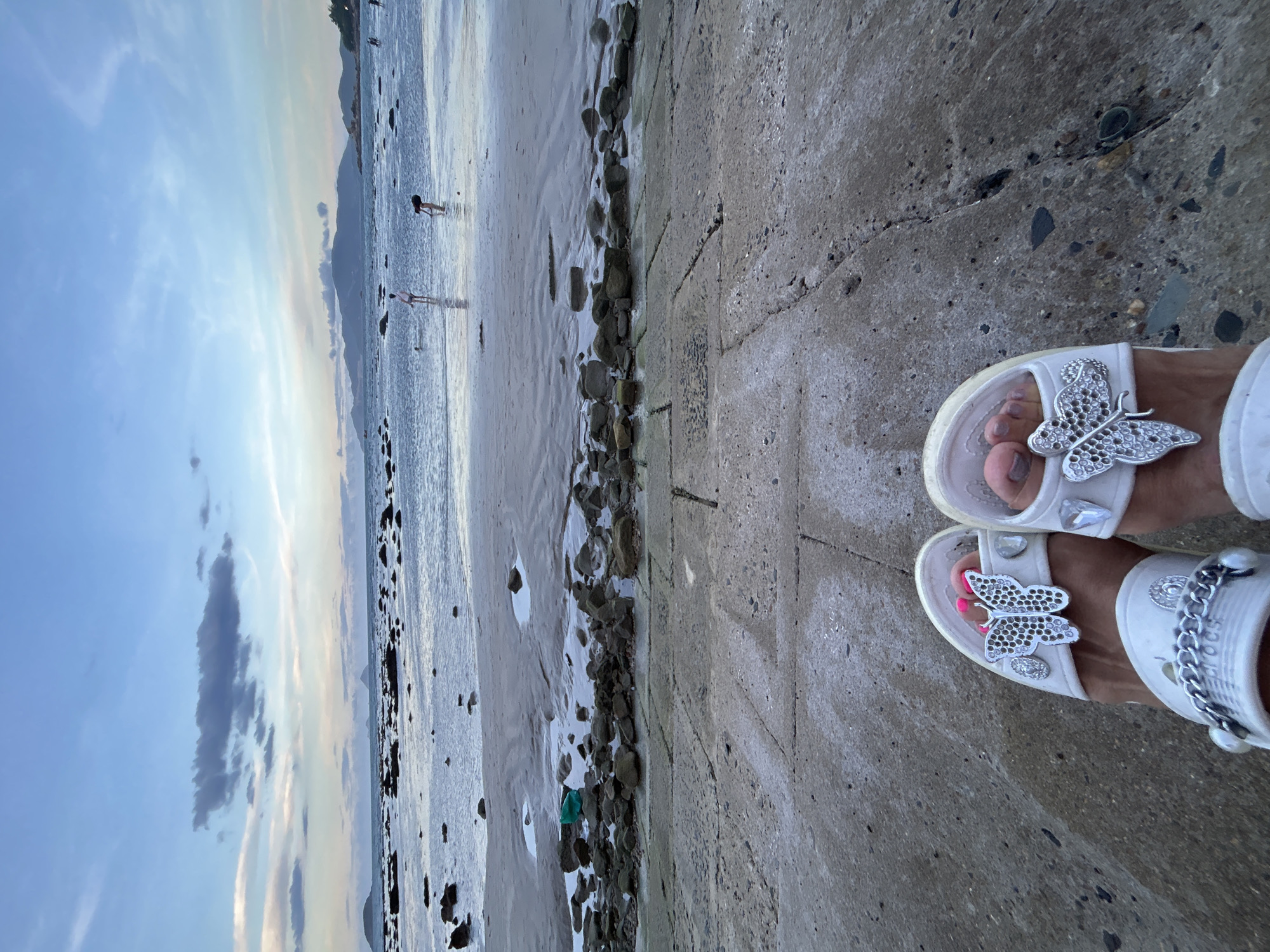A medium-shot, portrait-style, high-angle photograph shows two feet in white slide sandals with decorative butterfly appliques, positioned on a concrete walkway that is adjacent to a beach landscape. The sky is blue, with thin wisps of clouds scattered across the upper left of the image. The beach consists of gray sand and a body of water. Several people are seen in the distance, at the edge of the water. Along the beach, between the sand and concrete path, is a strip of stones and rocky formations. The concrete walkway is cracked and worn, with visible seams or divisions. The photograph was taken in Bắc Nha Trang Ward, Vietnam.