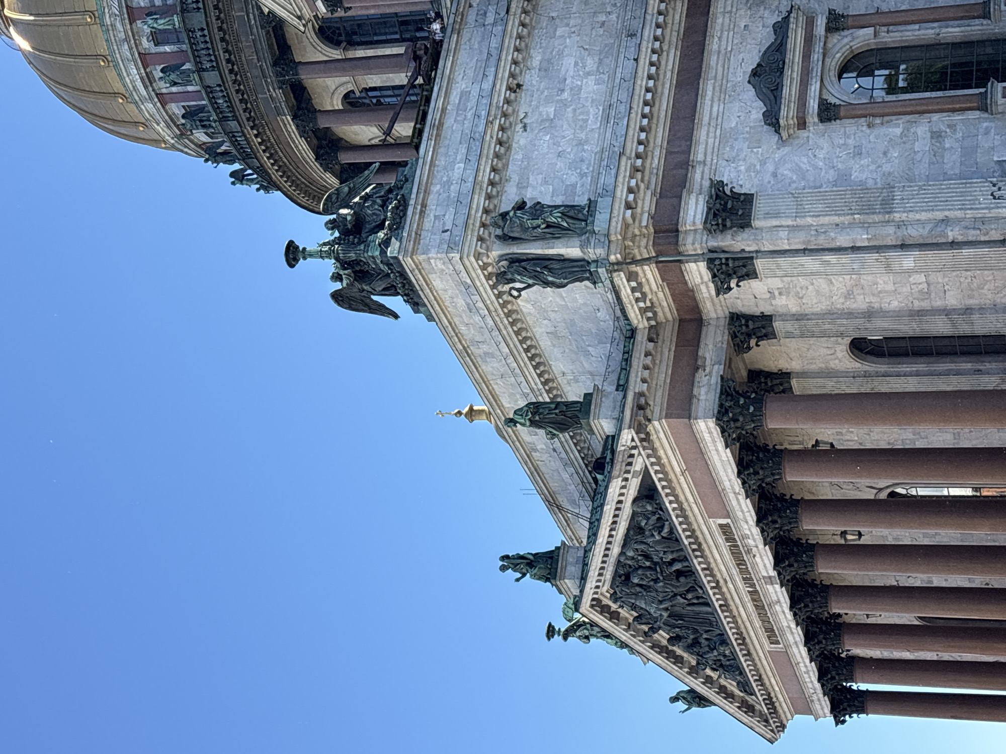 The image shows a close-up, low-angle view of the exterior of Saint Isaac's Cathedral in Saint Petersburg, Russia. The architecture is predominantly neoclassical, characterized by massive granite columns, intricate sculptures, and a gilded dome, parts of which are visible in the upper right corner. Several statues, including winged figures and robed individuals, adorn the roofline and cornices. The building's facade is constructed of marble with reddish-brown marble columns supporting the structure. The sky is a clear, bright blue, suggesting fair weather. No people are clearly identifiable in the scene.