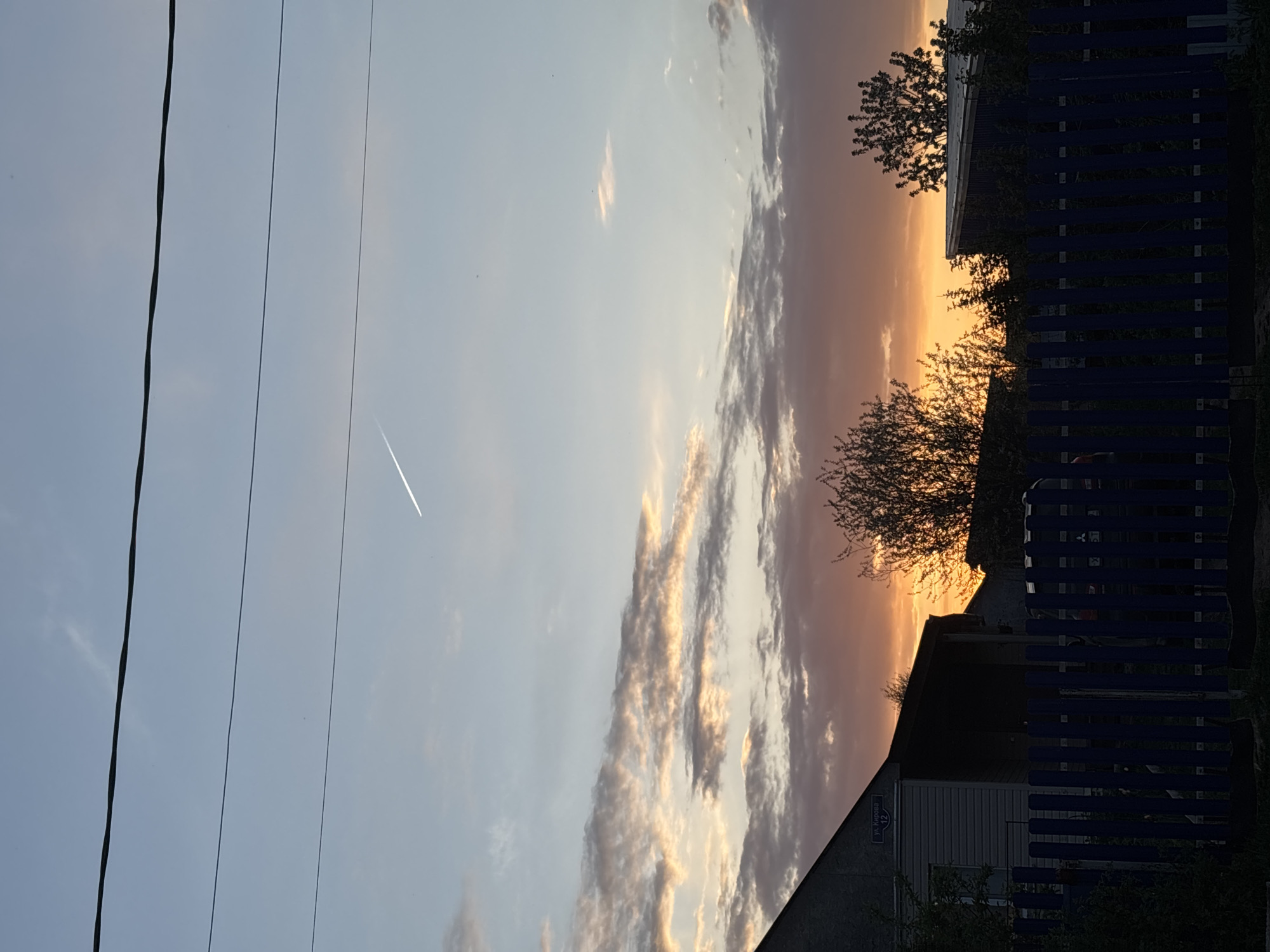 The image captures a twilight scene in Фоки, Russia, evidenced by the street sign "ул. Кирова 12" (Kirova Street 12) on a house. The sky transitions from light blue to orange and features elongated clouds. A contrail is visible in the upper-left of the sky. The scene is partially obscured by utility lines on the left. A wooden fence and silhouetted tree branches frame the scene on the right. A small house with a sloping roof is partially visible at the bottom right of the image.