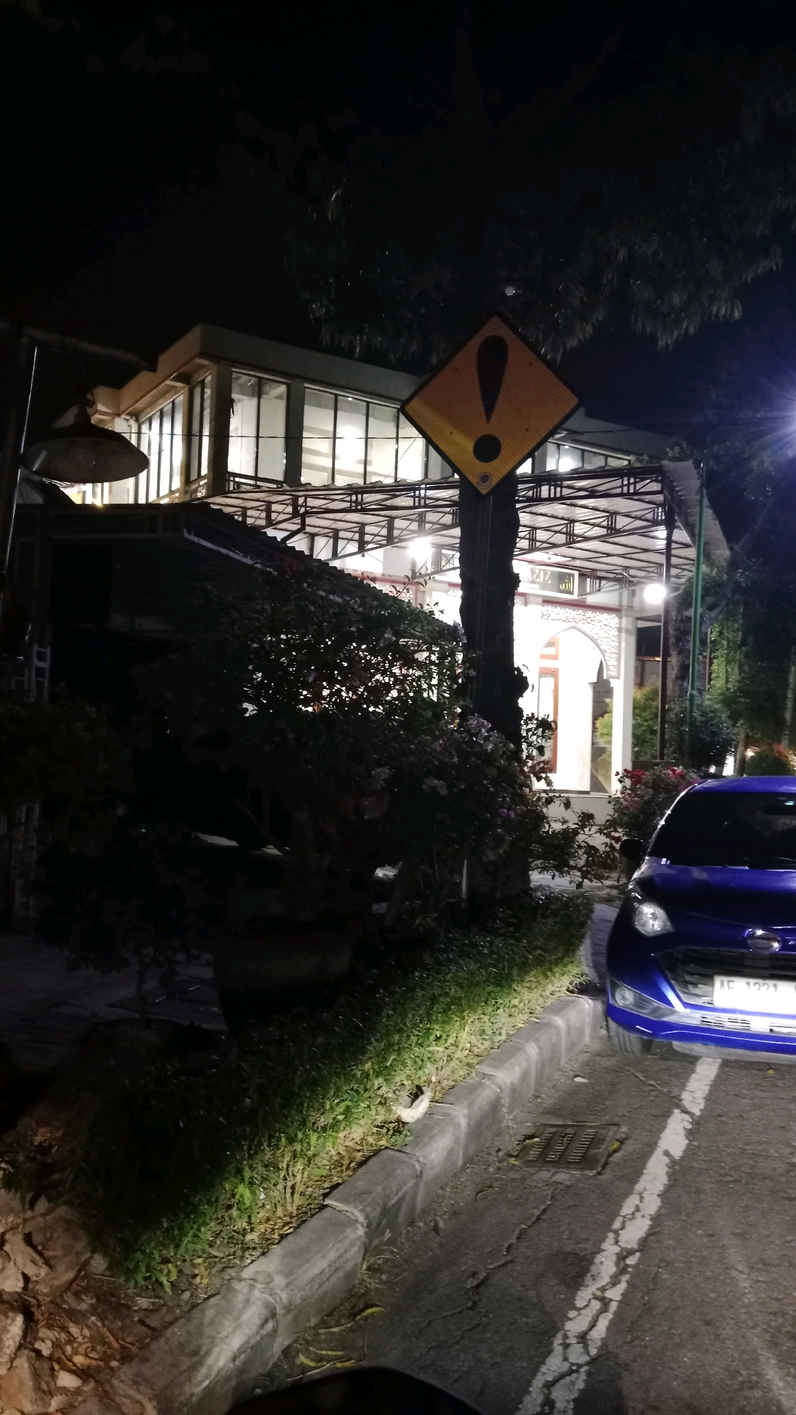 The image captures a nighttime scene on a street in Sukowinangun, Indonesia. A blue car is parked on the right side of the road, with its headlights on. The car's license plate reads "AE 1221 YN". Adjacent to the road is a sidewalk with a concrete curb, bordered by grass and vegetation. In the background, a modern building with large windows is illuminated from within. A yellow diamond-shaped warning sign with an exclamation mark is prominently displayed on a pole near the building. The overall atmosphere is dimly lit, with artificial lights from the building and a street lamp illuminating the scene. The building appears to be a public space or business, given its architecture and lighting.