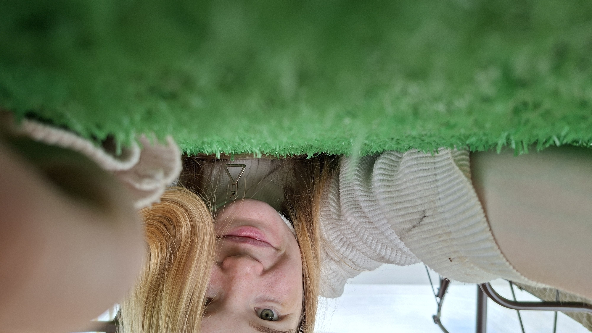 The image shows a young woman lying on artificial turf, looking at the camera. She has blonde hair and is wearing a white turtleneck sweater. Her right arm is extended towards the camera, and her hand is blurred. The background is out of focus and shows what appears to be an outdoor setting with some structure, possibly a balcony or a covered area. The lighting suggests it is daytime. The specific location in the city of Большой Букор, Russia cannot be determined from the image itself.