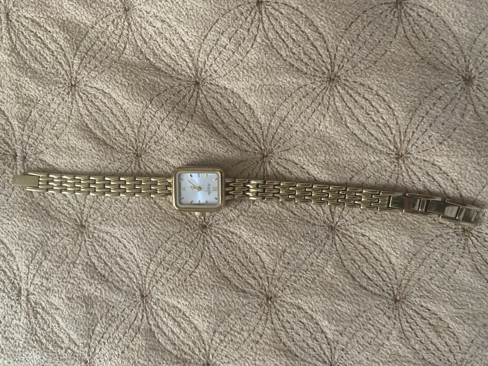 A gold-toned watch with a square face and a link band is laid diagonally on a textured, beige fabric surface. The watch face is light-colored with Roman numerals and thin, elegant hands. The brand name "sittric" is visible below the 12 o'clock position. The fabric has a repeating pattern of curved lines radiating from central points, creating a floral or sunburst-like effect. The image is a top-down view. No people are present in the image. The location of this image within the city of Kizilyurt, Russia, is unknown.