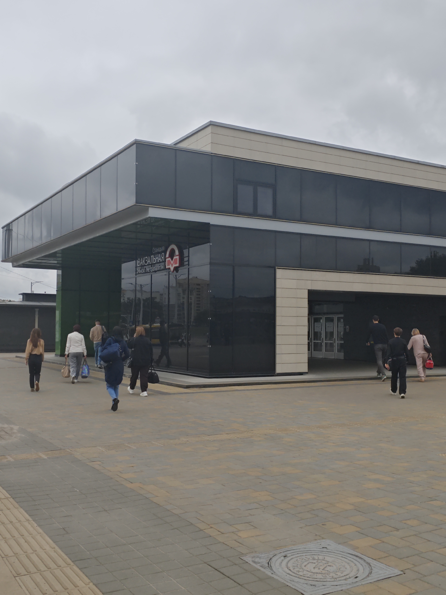 The image shows the entrance to the Vokzalnaya metro station in Minsk, Belarus. The building is modern with a glass facade and beige accents. Several people are entering or exiting the station. In the foreground, a man and a woman are walking away from the entrance, with the woman carrying a red bag. Another man in a dark top and grey pants is walking towards the entrance. Further to the left, a group of four people, two women and two men, are walking towards the entrance. The ground is paved with gray and beige rectangular stones, with yellow tactile paving visible. The sky is overcast.