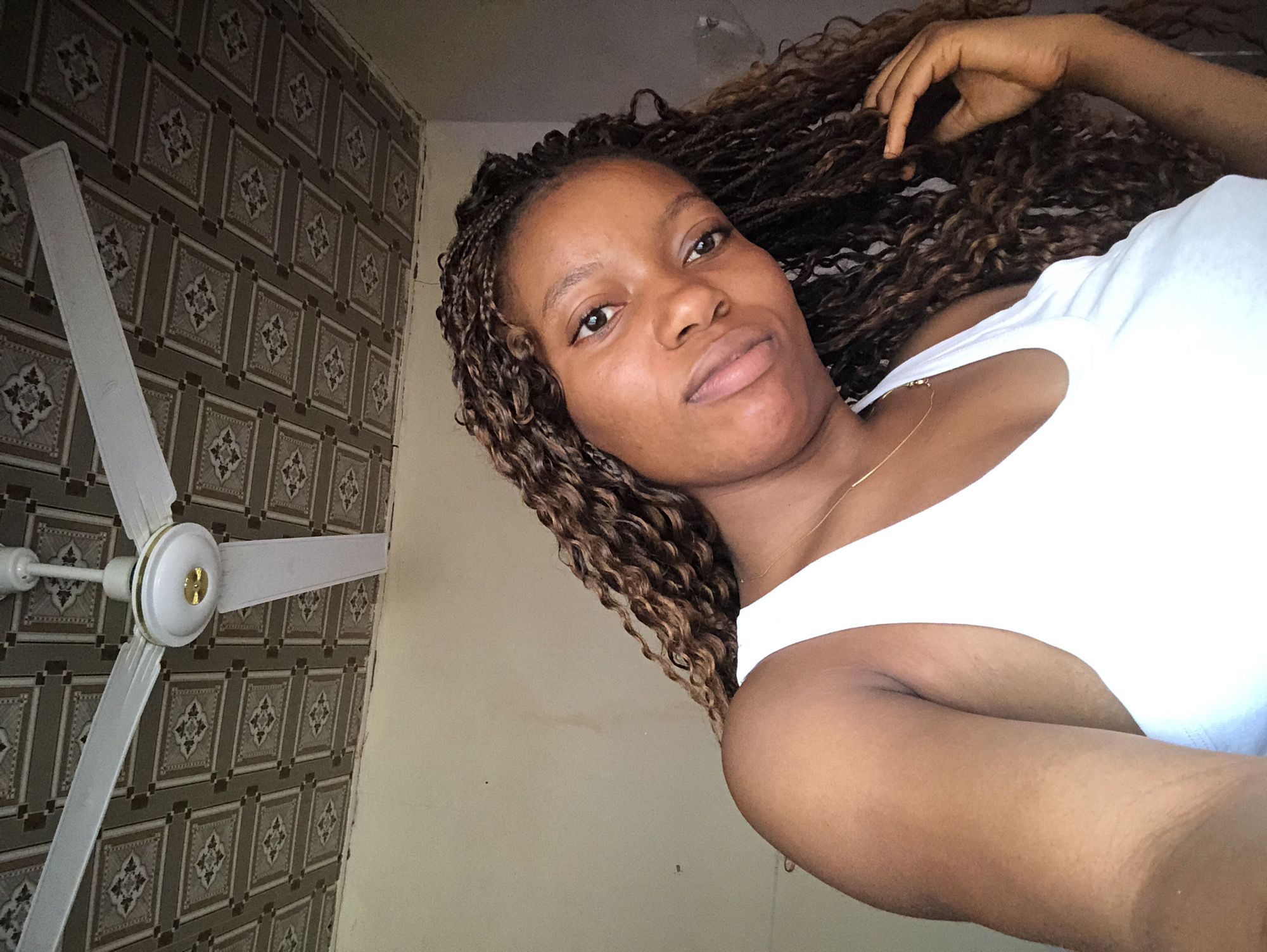 The image shows a woman with brown, braided hair, wearing a white tank top. She is facing the camera with a neutral expression. The background includes a wall with patterned wallpaper and a ceiling fan with white blades. The location is Kashere, Nigeria, based on contextual information.