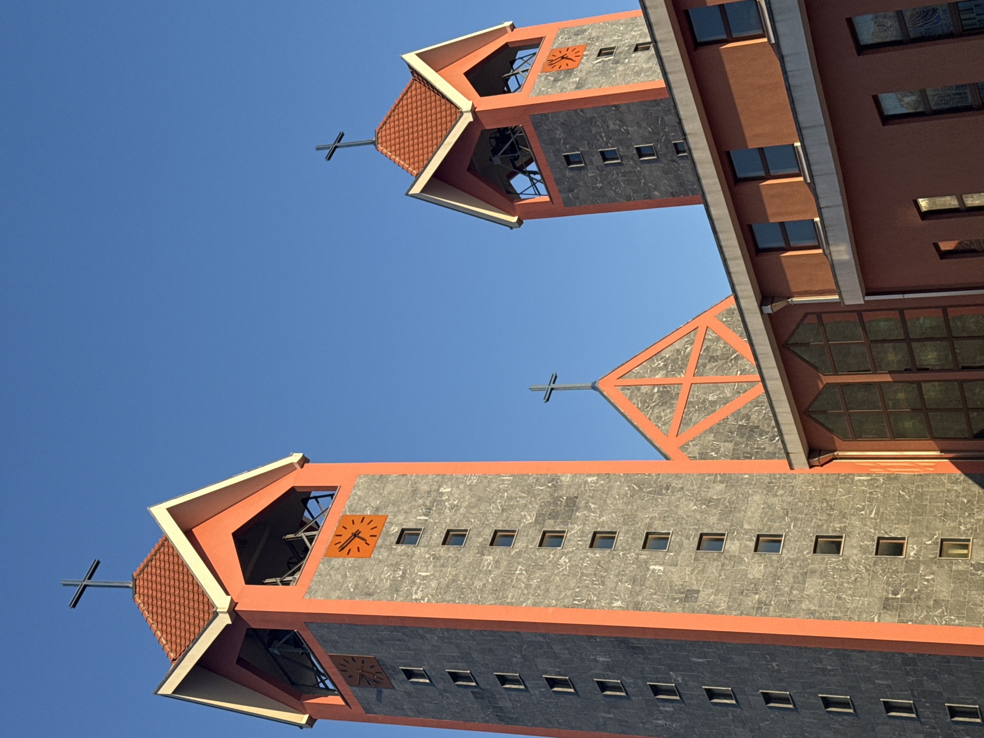 The image shows the Church of St. Peter the Apostle in Bar, Montenegro, viewed from a low angle, looking upwards towards the sky. The church is a modern design with two prominent towers. The towers feature grey tiled facades and orange trim. Each tower is topped with a cross. One tower has an exposed bell structure and a clock face. The sky is clear and blue.