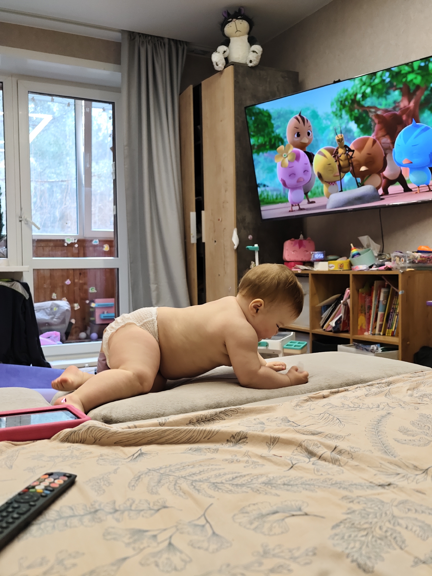 A baby wearing a diaper is lying on their stomach on a bed. A television is on in the background, showing a cartoon. The room appears to be a living room or bedroom with a window and curtains, a wardrobe, and a shelf with various items. A remote control and a pink tablet are on the bed. The scene is likely located in Chaikovsky, Russia.