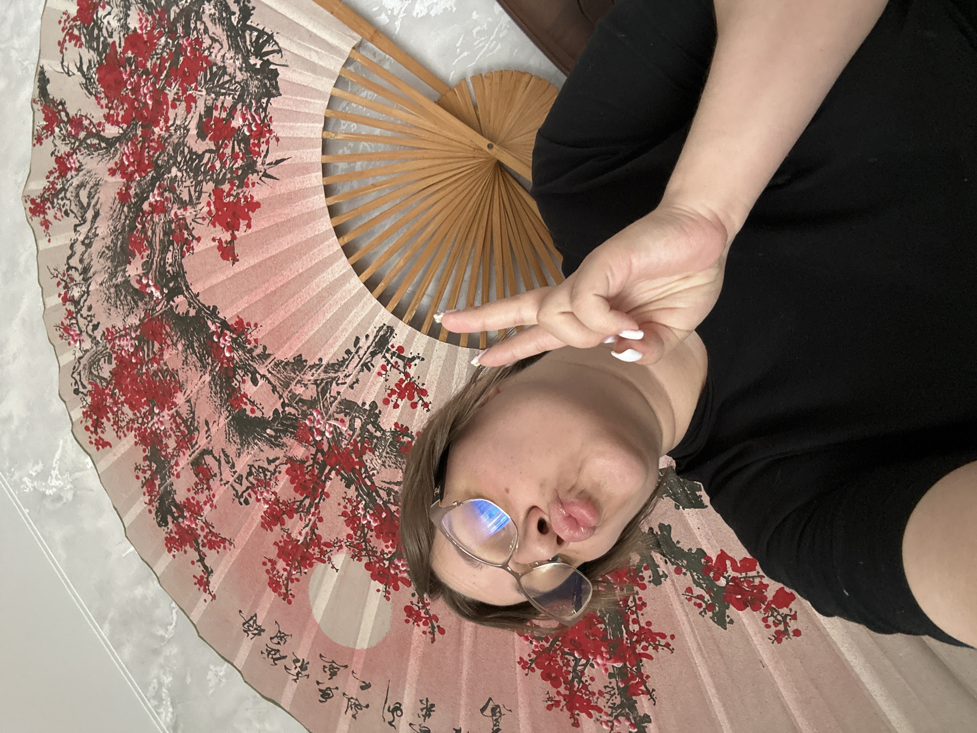 A person with glasses is lying on a large fan with red and black floral patterns on a light pink background. The person is wearing a black shirt and holding up a peace sign. The location is not discernible from the image.
