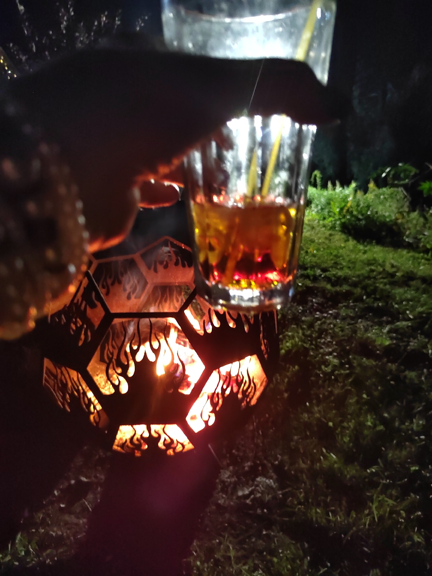 A person's hand holds a glass of amber-colored liquid with yellow straws, positioned over a hexagonal metal fire pit with flame cutouts. The fire pit contains a bright, glowing fire. The background shows dark foliage and grass, suggesting an outdoor evening setting. The image is dimly lit, with the fire and the liquid in the glass being primary light sources.