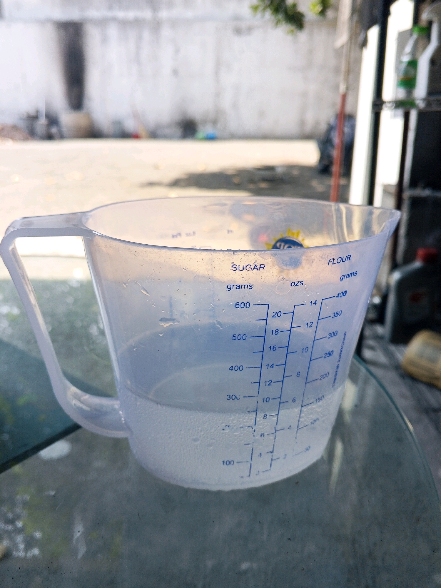 In Condongcatur, Indonesia, a clear plastic measuring cup, filled with a clear liquid and containing small bubbles, sits on a glass table. The cup has a handle and markings for "SUGAR" in grams and "FLOUR" in grams and ounces. The background is blurred, showing a concrete wall, outdoor ground, and some greenery. The lighting suggests it is daytime.