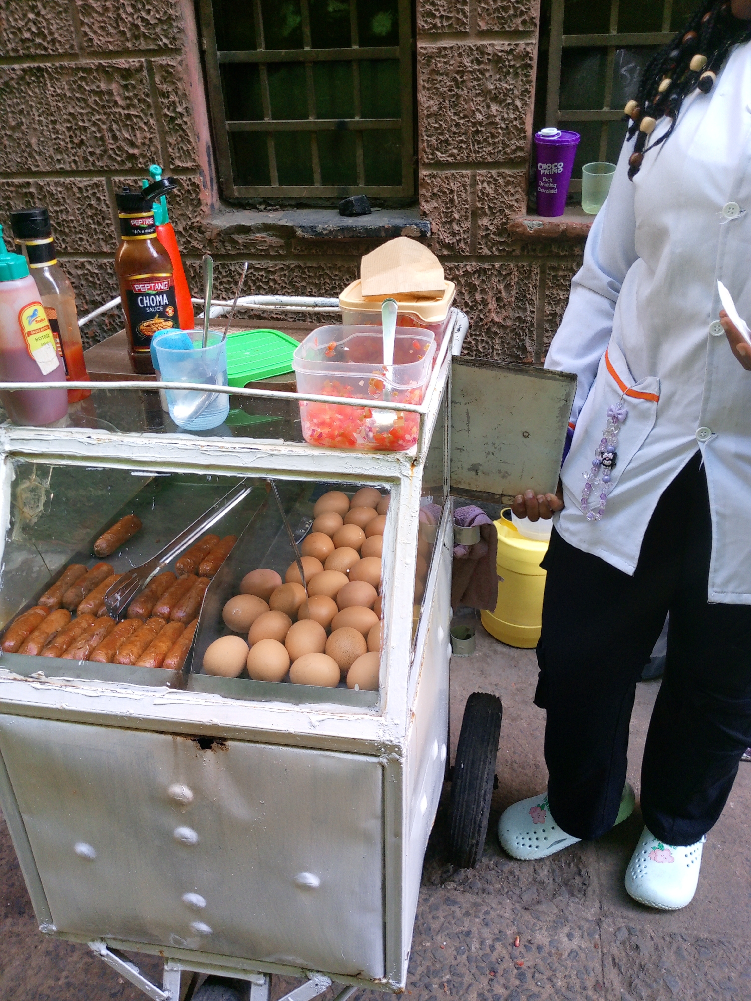 The image depicts an outdoor street food vending operation located against a textured reddish-brown brick building in Nairobi, Kenya. A partially visible individual stands to the right of a white metal food cart. This person wears a white long-sleeved chef's shirt with orange trim on the pocket, black trousers, and light green clogs with a floral design. Their dark braided hair, adorned with light-colored wooden beads, is visible over their right shoulder. Their right hand holds a small white utensil or object.
The food cart features a glass display section. The top surface of the cart holds several items: two bottles of sauce (one labeled "PEPTANG CHOMA SAUCE," the other a generic red sauce), a light blue plastic cup containing metal spoons, and a clear plastic container filled with diced red produce, consistent with tomatoes or salsa. The main display area below is divided into two sections by glass. The left section contains multiple cooked sausages or hotdogs, with a pair of metal tongs resting among them. The right section holds dozens of raw brown eggs. The lower metal panel of the food cart shows signs of wear, including dents and a small area of rust.
In the background, the brick building features two windows protected by dark metal bars. On the window ledge, a purple cup or thermos labeled "Choco Primo" and a clear glass are present. A yellow plastic container is visible on the ground directly behind the food cart. The ground surface is dark and paved.