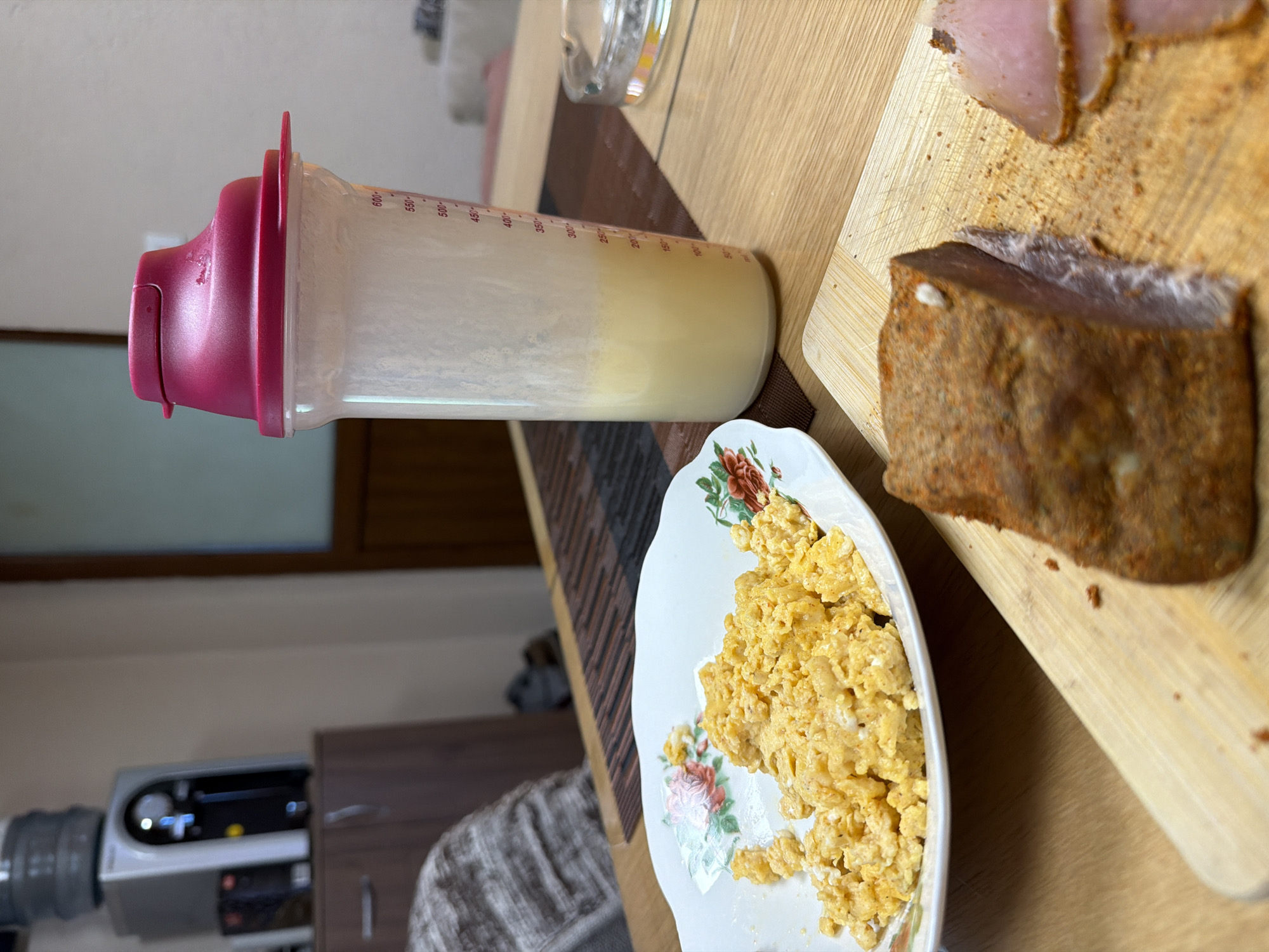 The image presents a scene on a wooden table. A plastic shaker bottle, filled partially with a yellowish liquid, sits horizontally. A plate of scrambled eggs, decorated with floral patterns, is placed next to the bottle. Adjacent to the eggs, on a wooden cutting board, are slices of meat, likely ham and another brown-crusted piece of meat. In the background, partial views of a water cooler and a brown cabinet can be seen. The image orientation suggests it was taken in portrait mode. Location details of Panagyurishte, Bulgaria are not evident from the image.