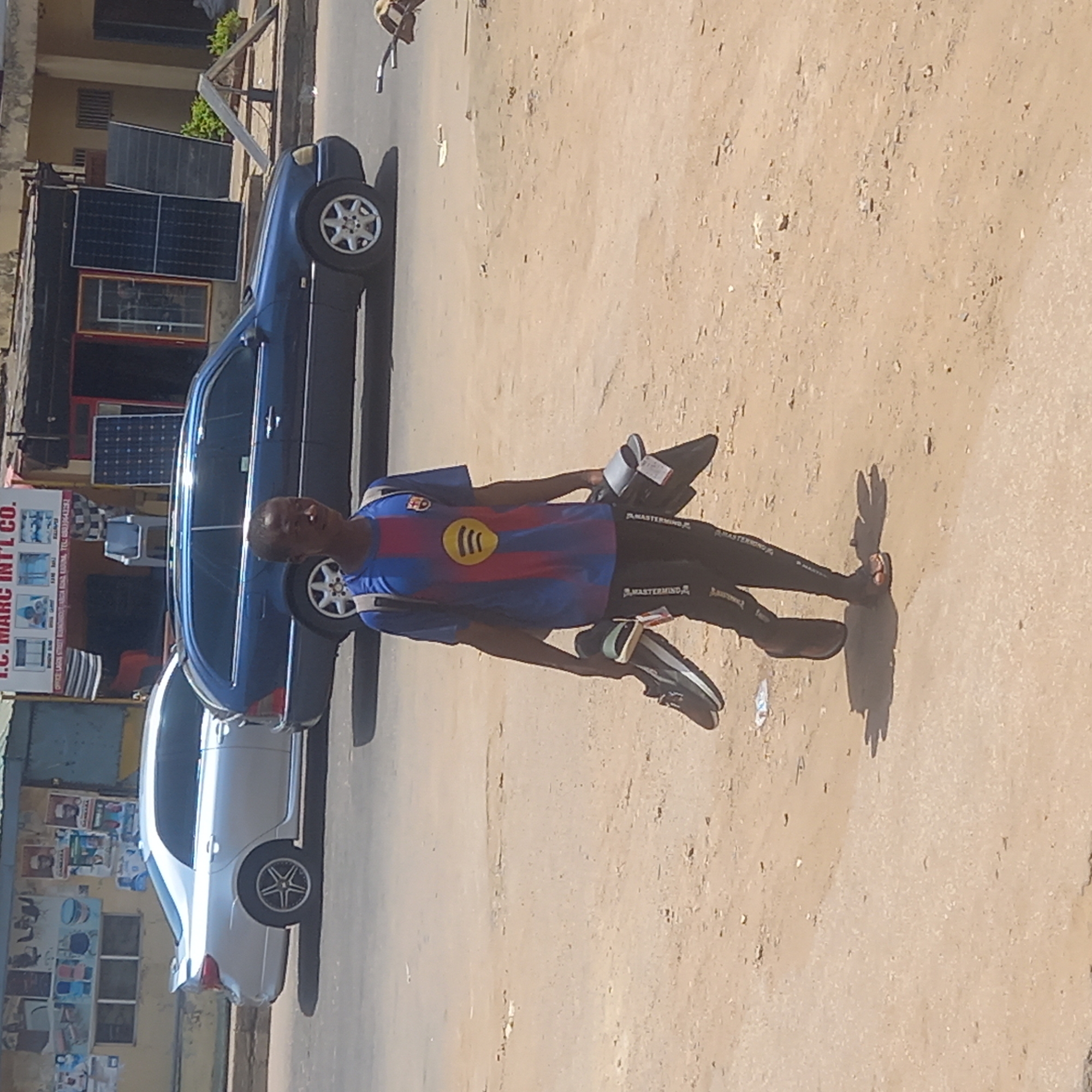 A man stands on a dusty street in Dadi Riba, Nigeria, holding a pair of shoes in each hand. He is wearing a blue and red striped shirt with a yellow design, dark pants with the word "MASTERMIND" printed on them, and sandals. A blue sedan and a white car are parked alongside the street. A building with a sign that reads "T.C. MARC INT'L CO." is visible in the background, along with solar panels. The street is unpaved.