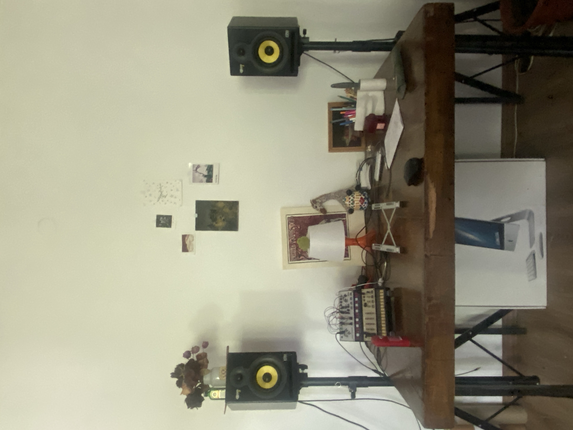 The image shows a section of a room interior. A wooden desk stands perpendicular to a white wall. Two studio monitor speakers are mounted on stands on either side of the desk. The top speaker is positioned higher than the bottom one. Various items are placed on the desk, including a lamp with a white lampshade, a modular synthesizer, pencils in a container, and a framed picture. Several small images are attached to the wall. A large white box with an image of an Apple product is also on the desk. There is no evidence to suggest the location is Шебунино, Russia, or any information about people, actions, or interactions in the image.